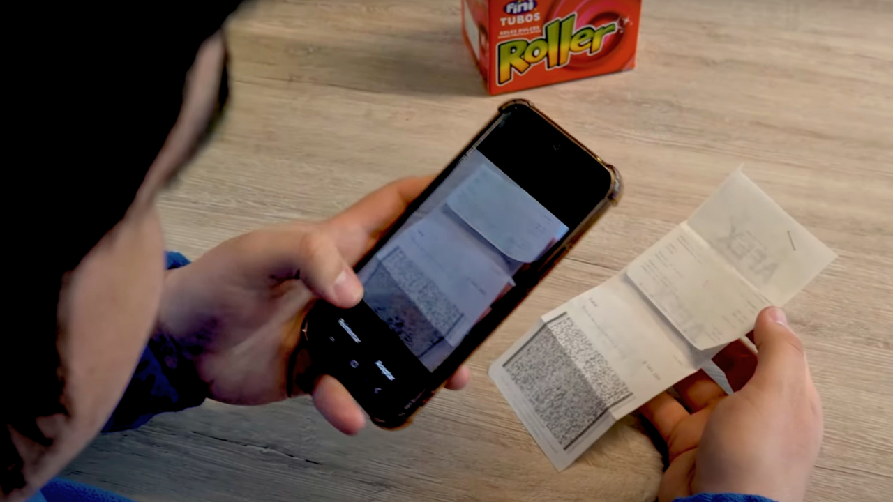 Persona escaneando recibos físicos con su smartphone sobre una mesa, en un entorno informal con productos de la marca Fini visibles al fondo, destacando procesos de registro de viáticos.