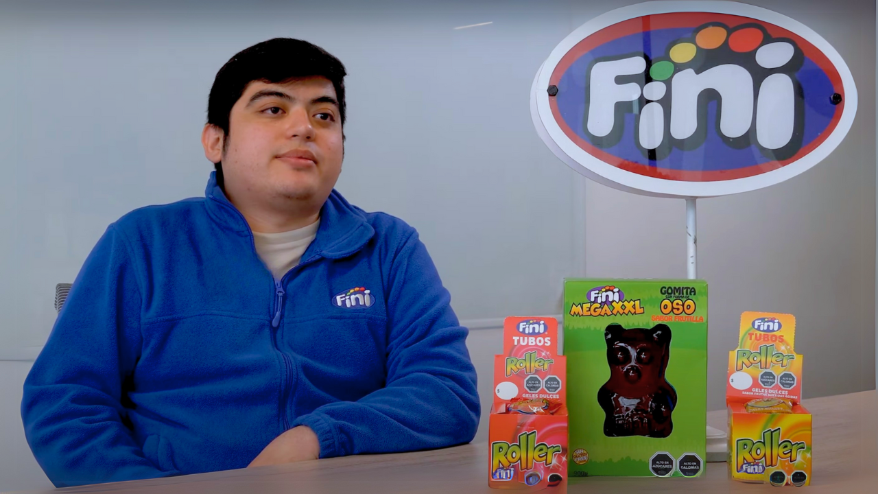 Colaborador de Fini vestido con sudadera azul con logotipo, sentado frente a productos de la marca y un cartel corporativo, en un ambiente de oficina durante una entrevista.