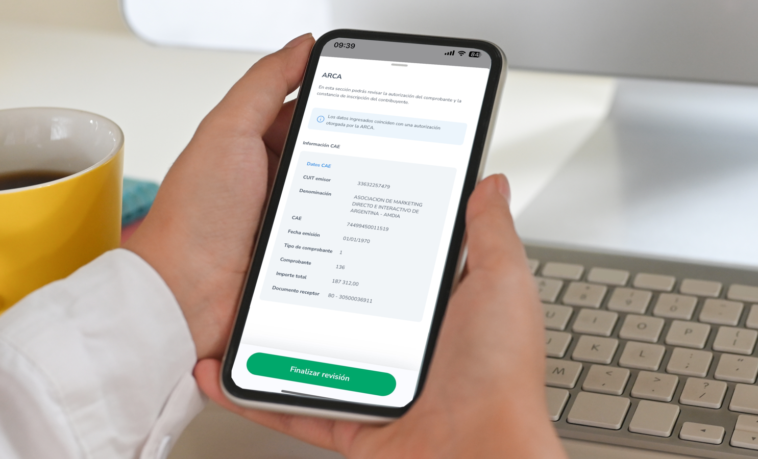 Persona sosteniendo un teléfono móvil con la aplicación ARCA abierta, mostrando la validación de un comprobante con datos de CUIT y CAE. En el fondo, un teclado de computadora y una taza de café amarilla sobre un escritorio