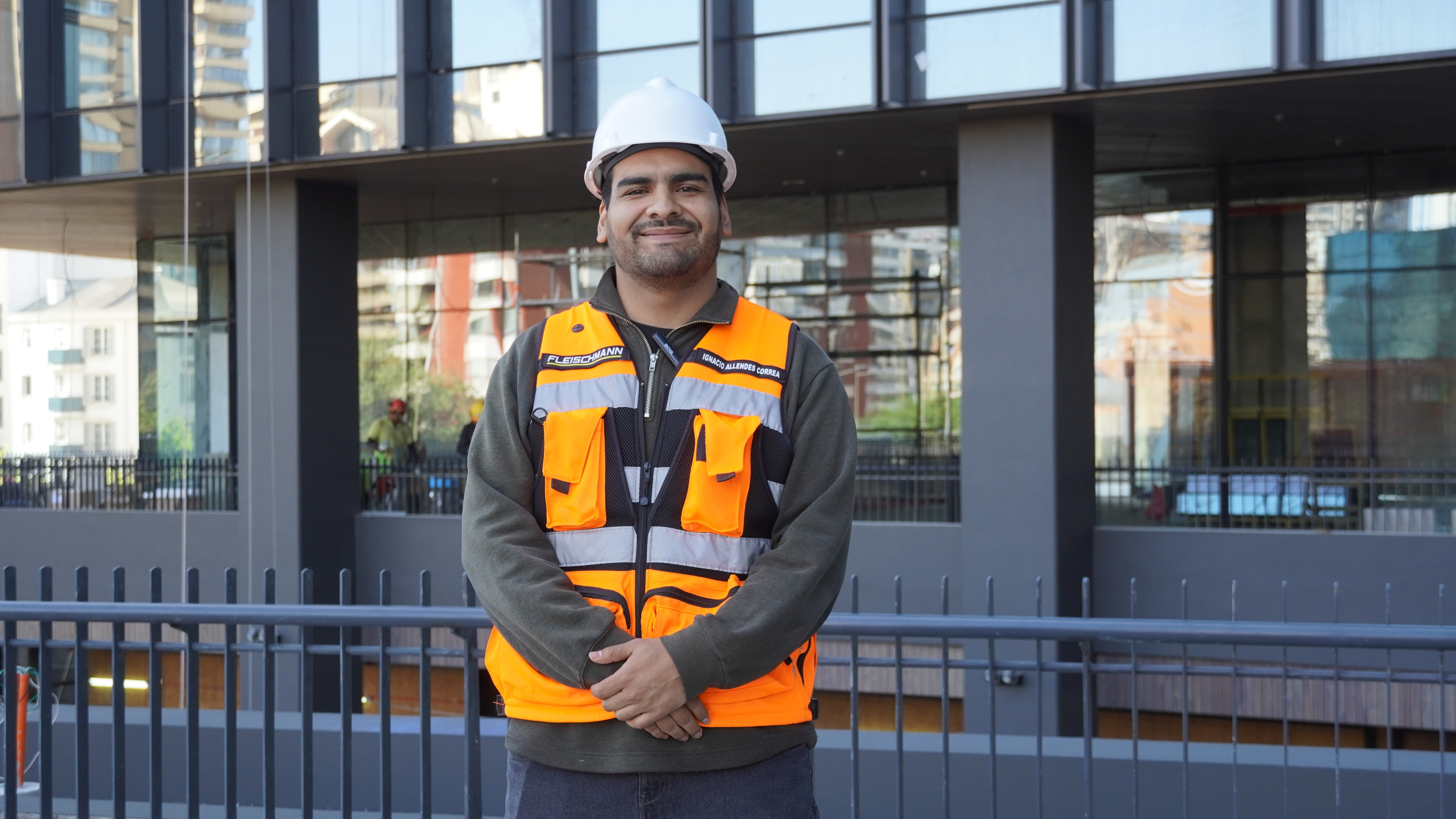 Empleado de Fleischmann S.A. posando frente a un edificio moderno, con casco blanco y chaleco reflectante anaranjado, representando digitalización en proyectos de construcción