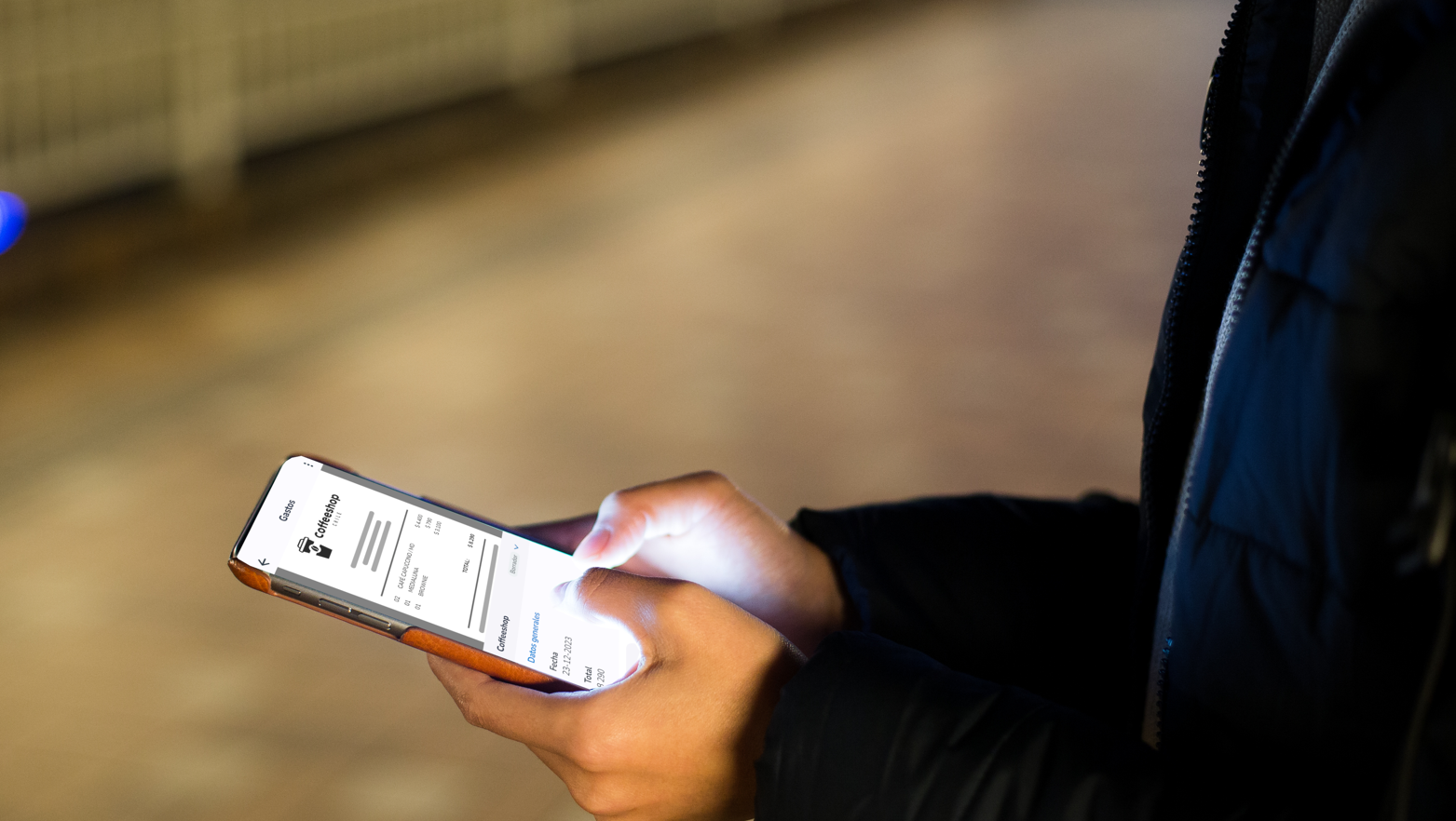 Persona usando un teléfono móvil en una estación de metro o un entorno oscuro, con la pantalla iluminada que muestra detalles de una factura o informe de gastos.