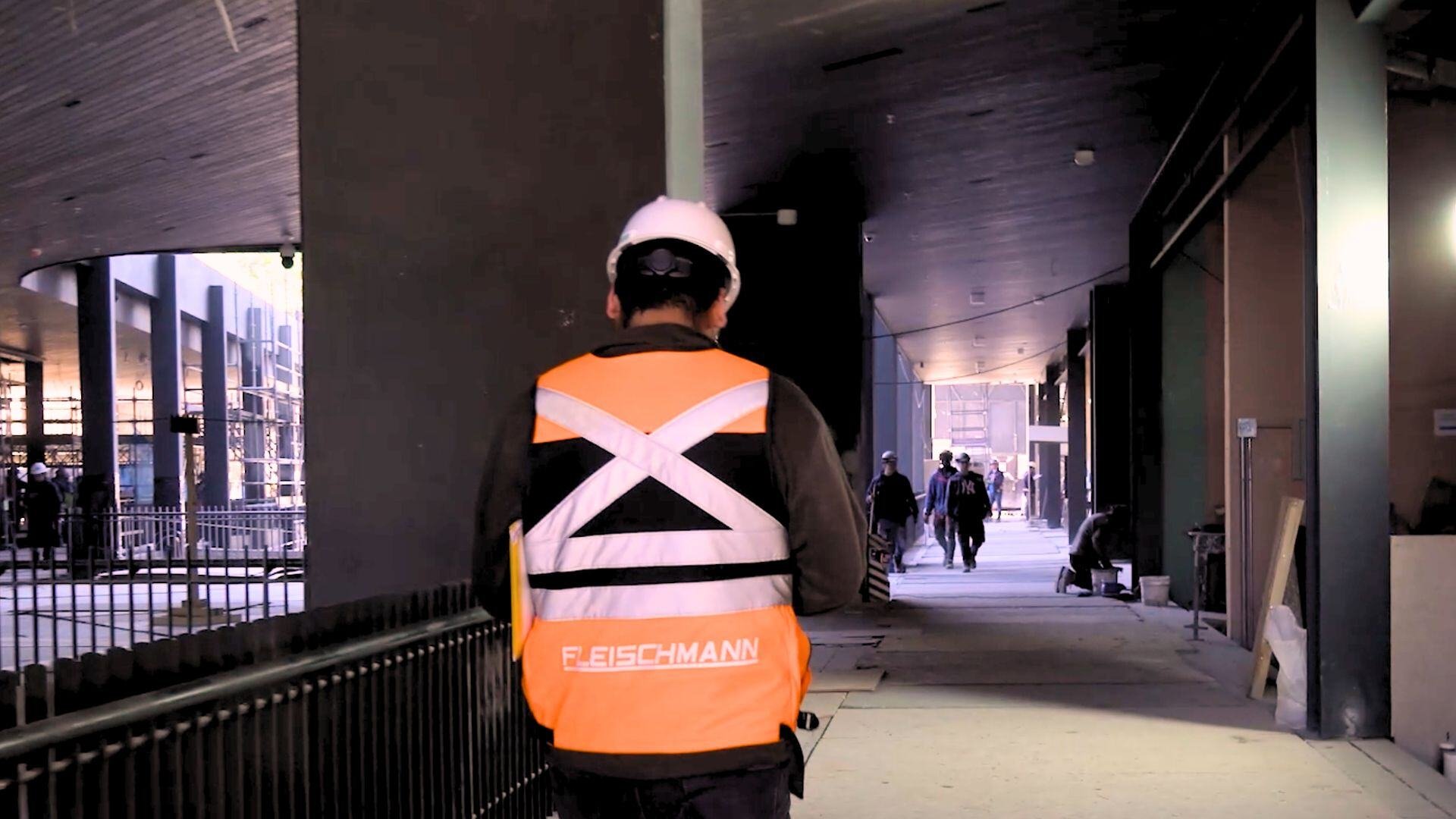 Trabajador de construcción de espaldas, usando casco blanco y chaleco reflectante de Fleischmann S.A., caminando por un pasillo de obra con iluminación tenue.