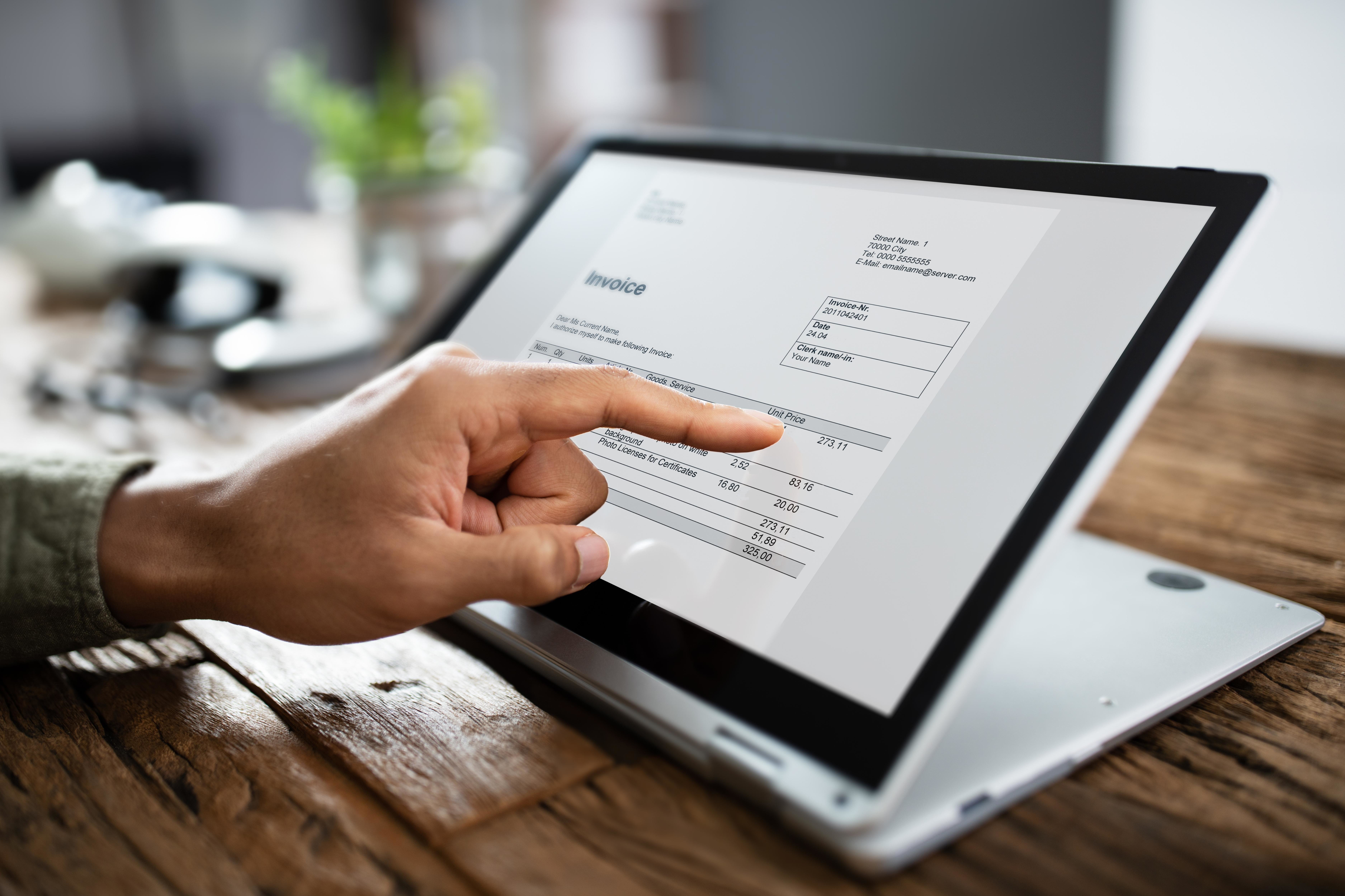 Persona señalando con el dedo detalles de una factura en una pantalla de laptop, con el fondo desenfocado y un ambiente de oficina cálido y ordenado.