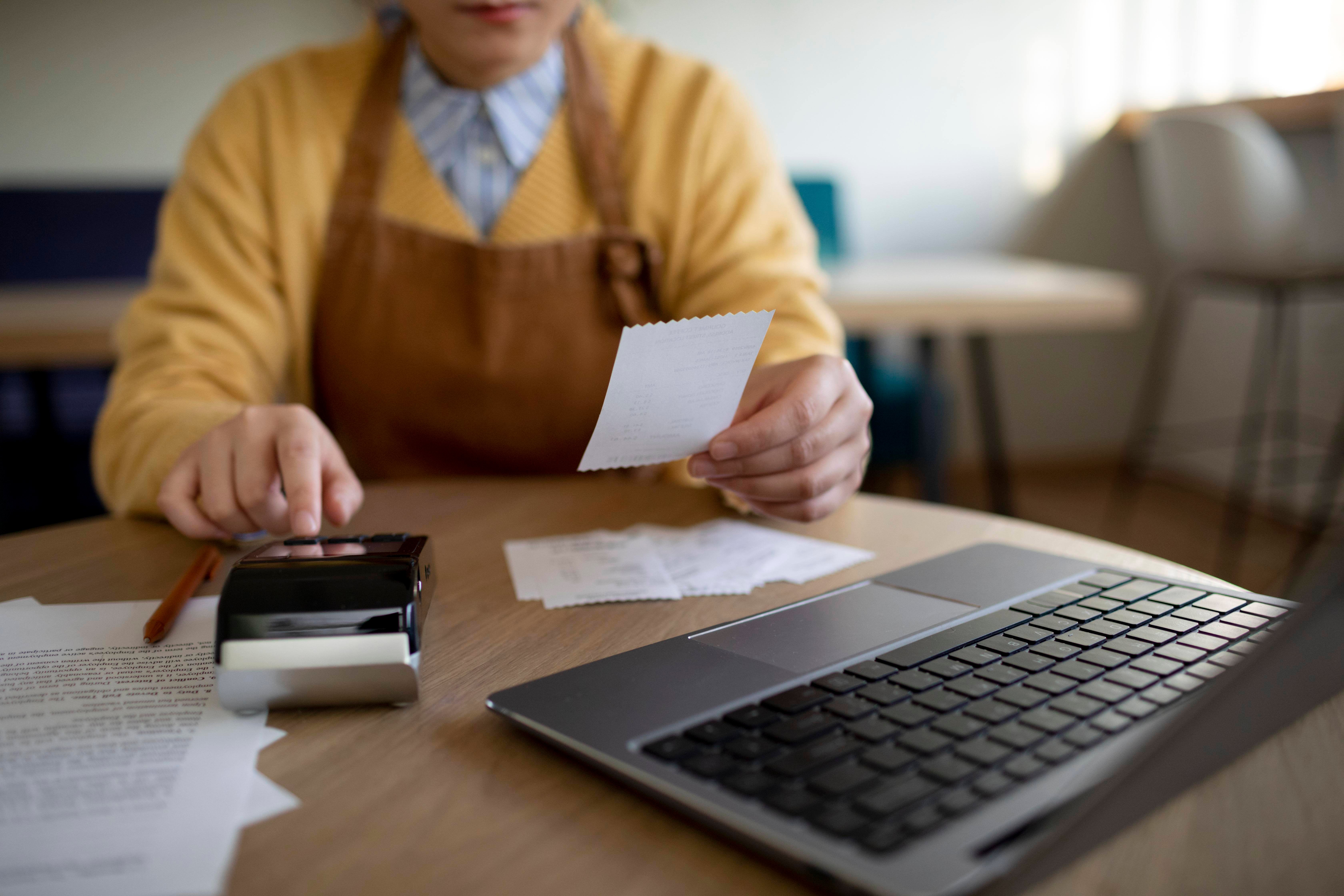 Persona con un recibo en la mano mientras utiliza una calculadora y una laptop, organizando sus gastos y documentos."