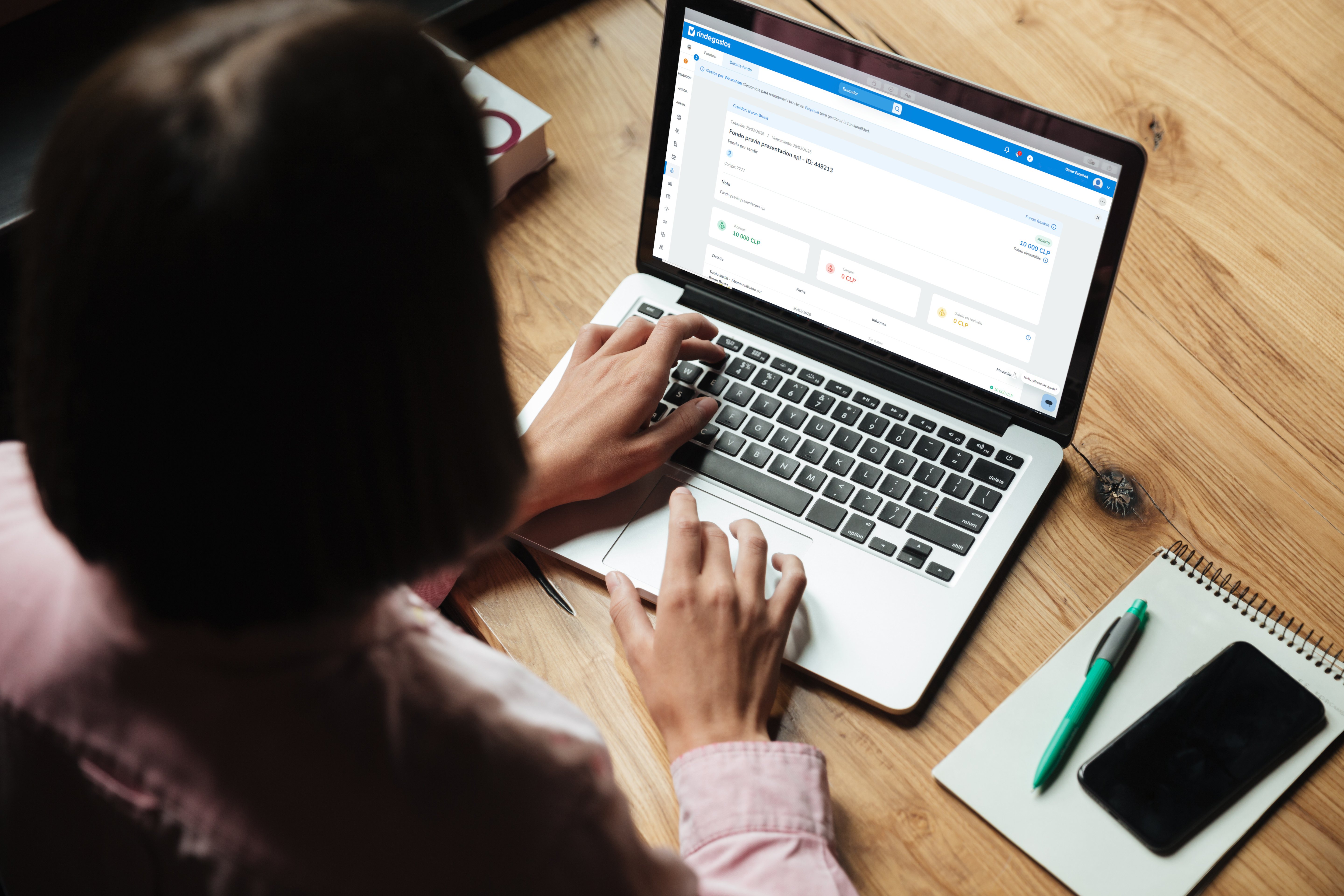 Persona usando una laptop con la plataforma de Rindegastos abierta en la pantalla. Se observa un escritorio de madera con una libreta, un bolígrafo verde y un teléfono móvil al lado.