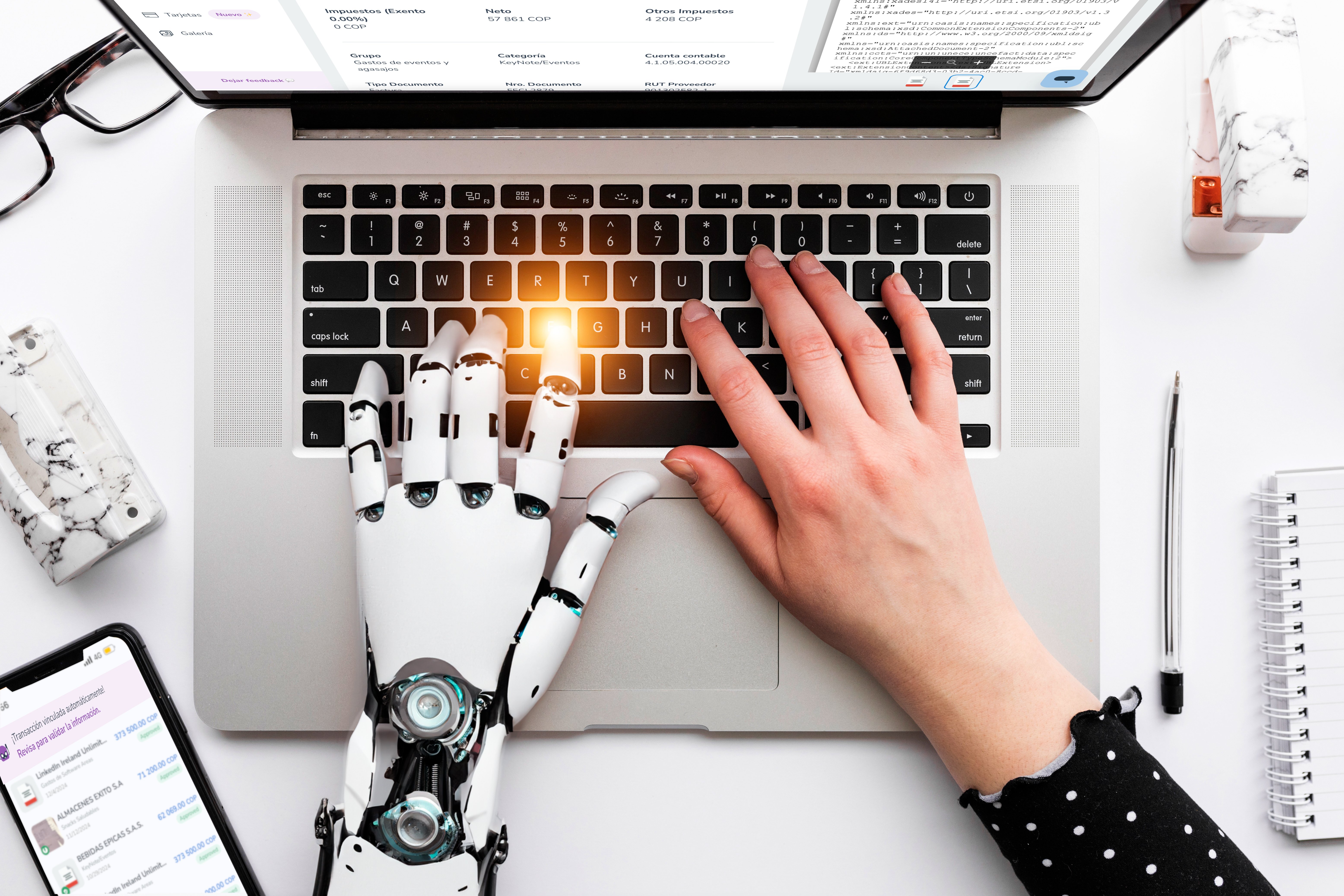  Mano humana y brazo robótico tecleando sobre una computadora portátil, con un teléfono móvil y otros objetos de oficina alrededor