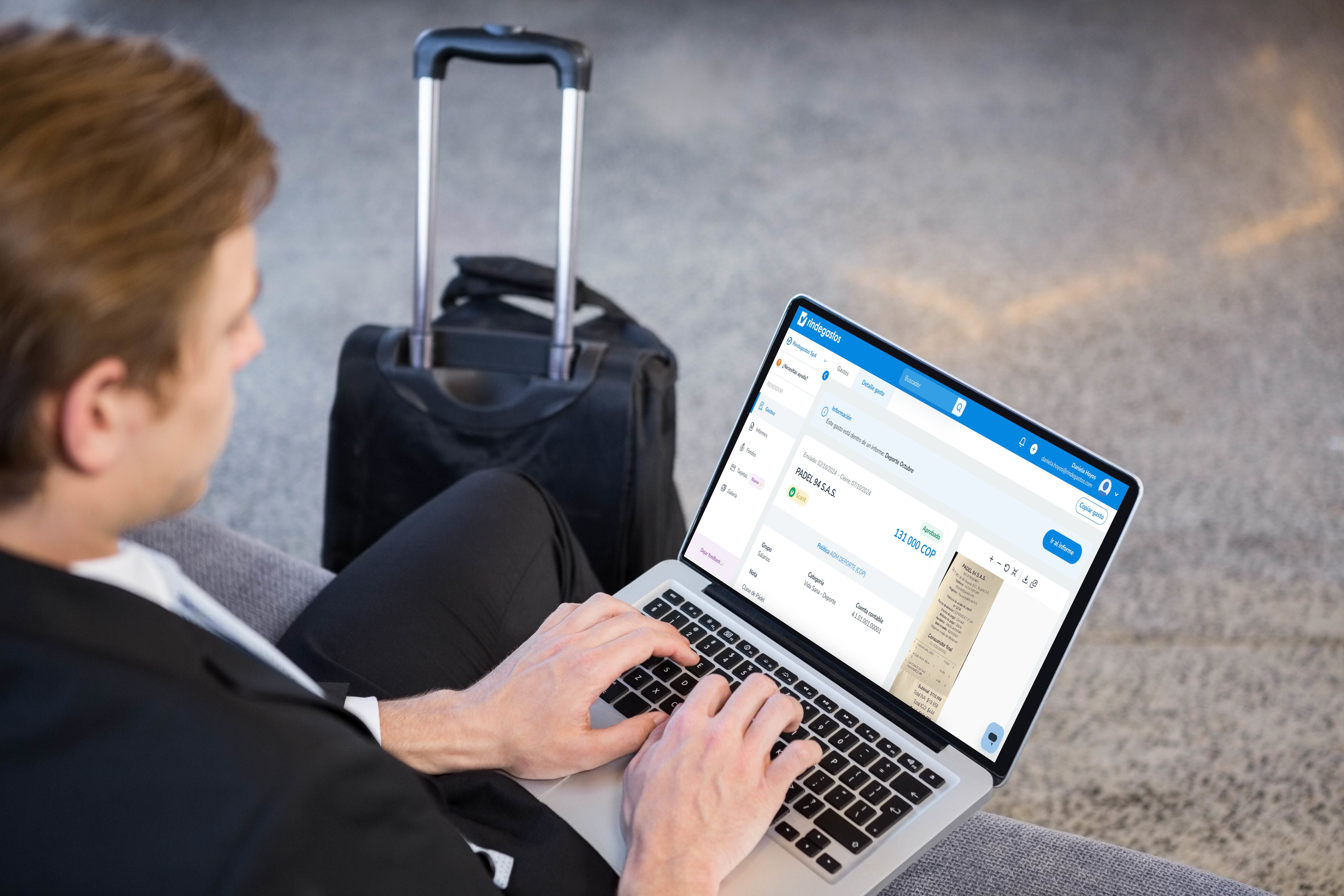 Persona de negocios utilizando una computadora portátil mientras está sentada en una sala de aeropuerto, con una maleta al fondo. La pantalla muestra Rindegastos