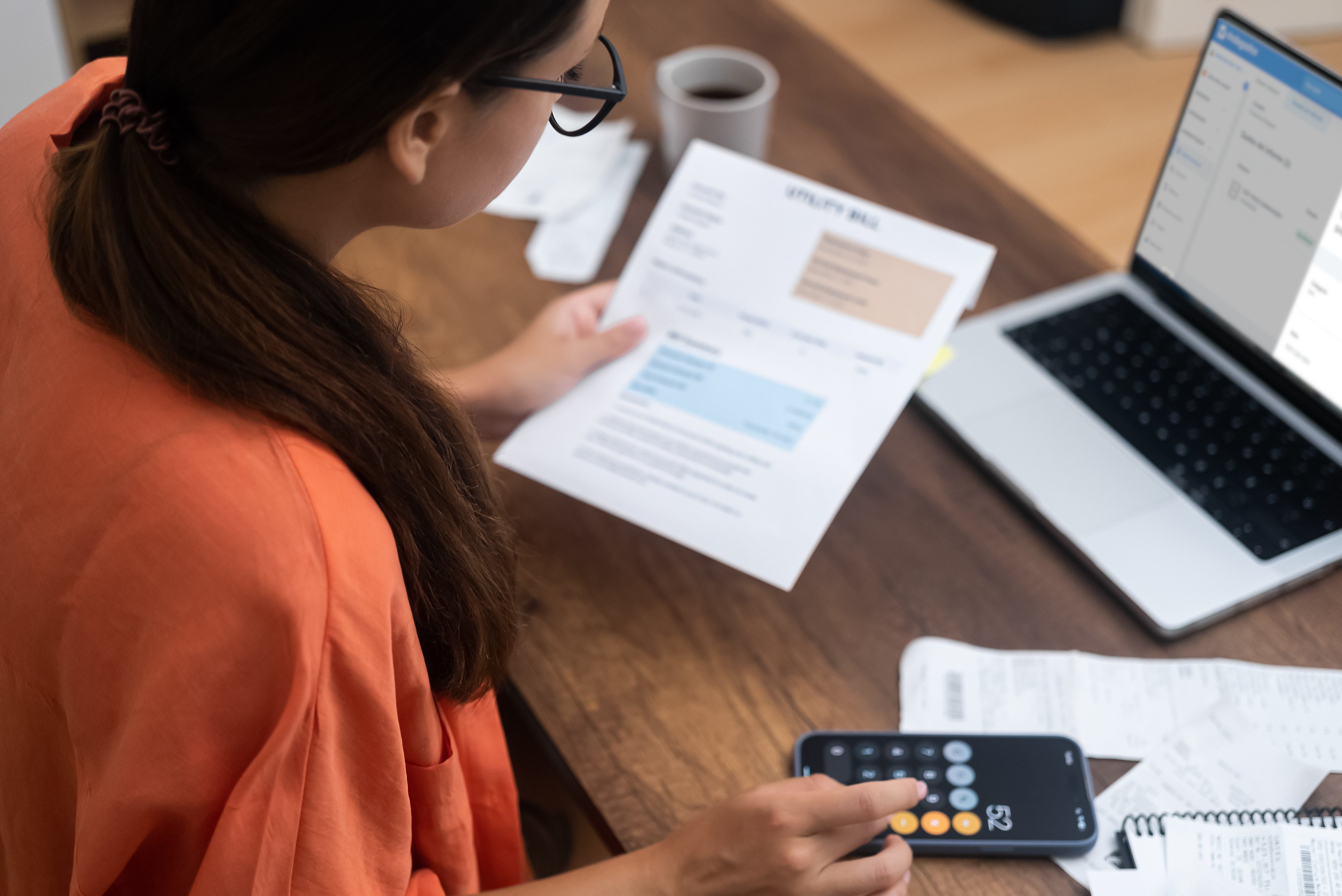  Mujer con gafas revisando una factura de servicios mientras utiliza una calculadora y su laptop en casa, organizando comprobantes sobre la mesa como parte de la gestión de gastos personales o empresariales.