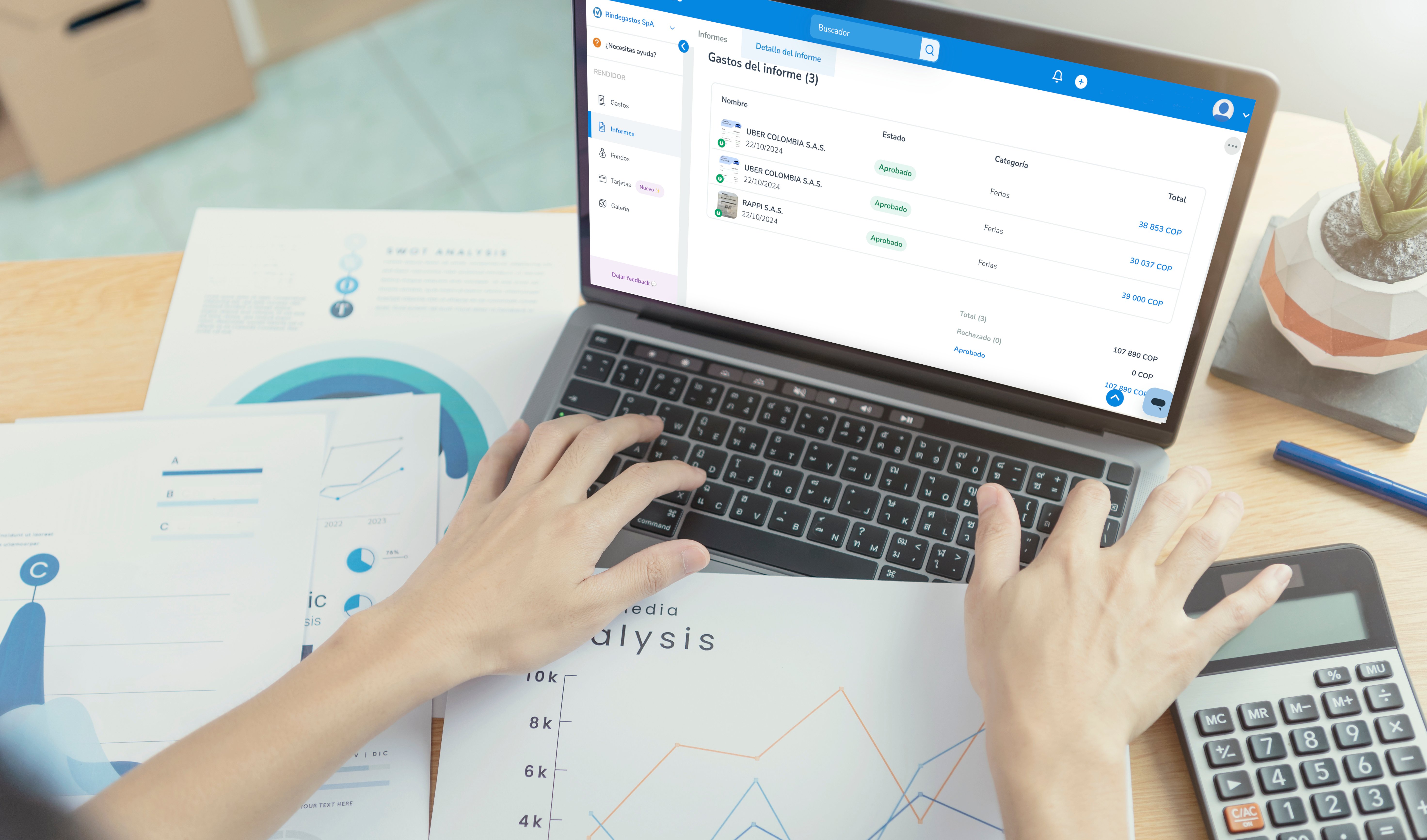 Persona trabajando en una laptop, con documentos de análisis de gráficos y datos financieros sobre la mesa, mientras la pantalla de la laptop muestra la plataforma de Rindegastos con un informe de gastos.