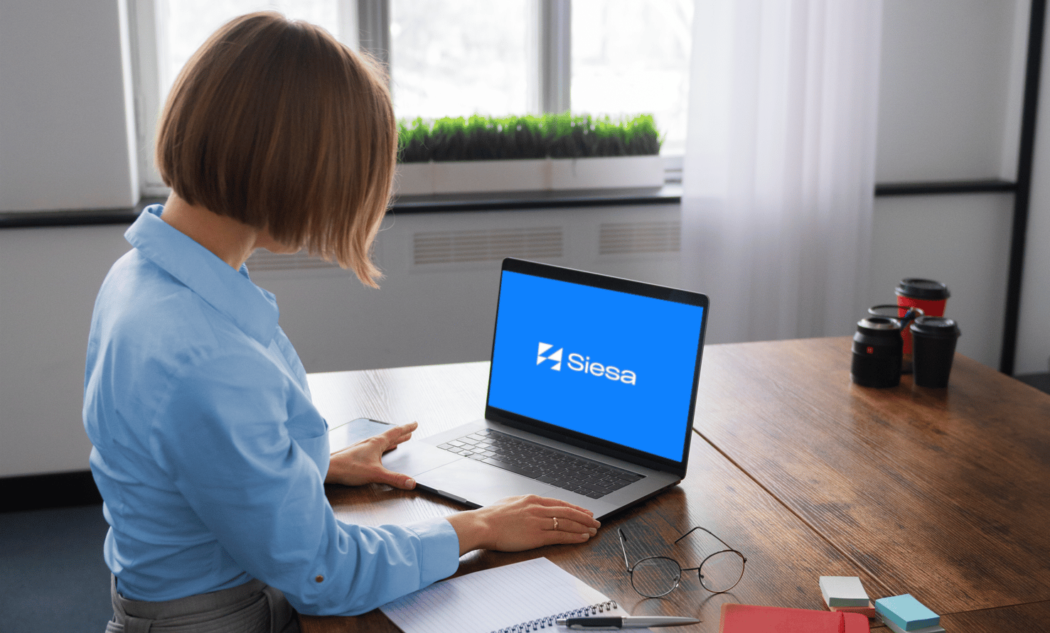 Profesional trabajando con el sistema Siesa en su laptop dentro de una oficina moderna, con tazas de café y lentes sobre la mesa.