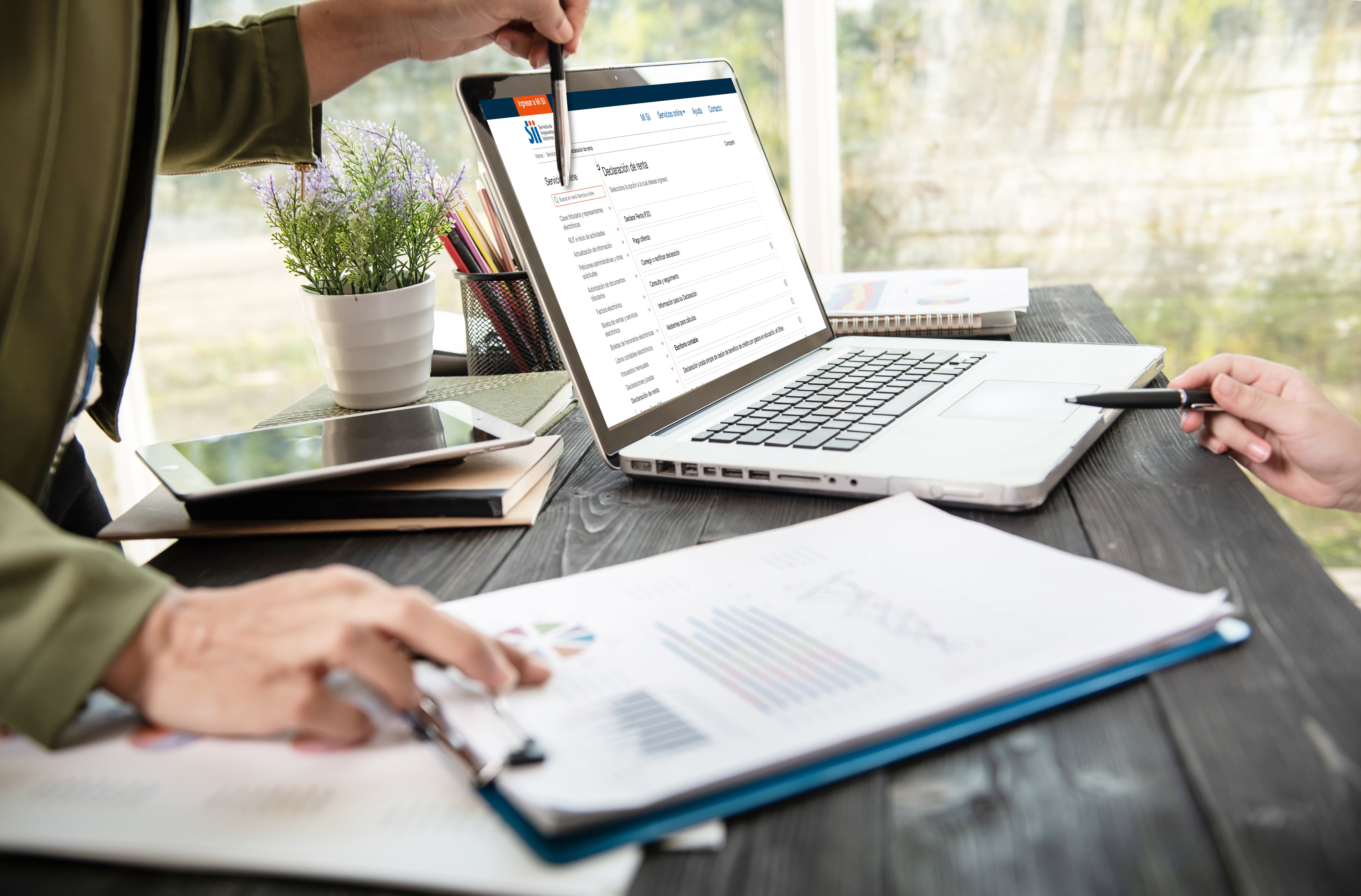 Personas trabajando frente a un computador con el sitio web del SII abierto, revisando información tributaria. En la mesa hay gráficos, una tablet y una planta decorativa.