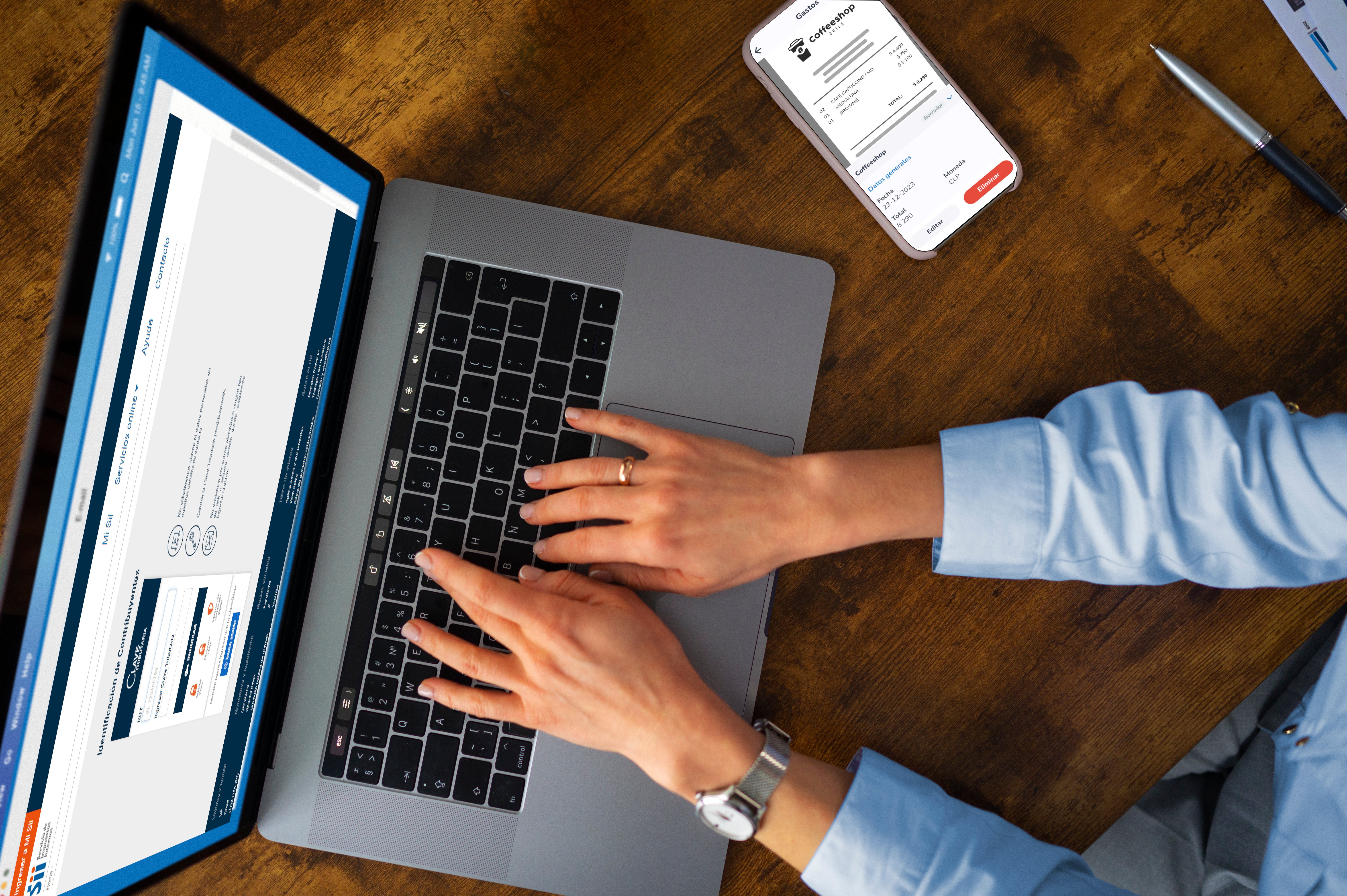 SIIPersona utilizando una laptop mientras revisa una factura en su teléfono móvil, con una mano tocando la pantalla del móvil y la otra sobre el teclado de la laptop.