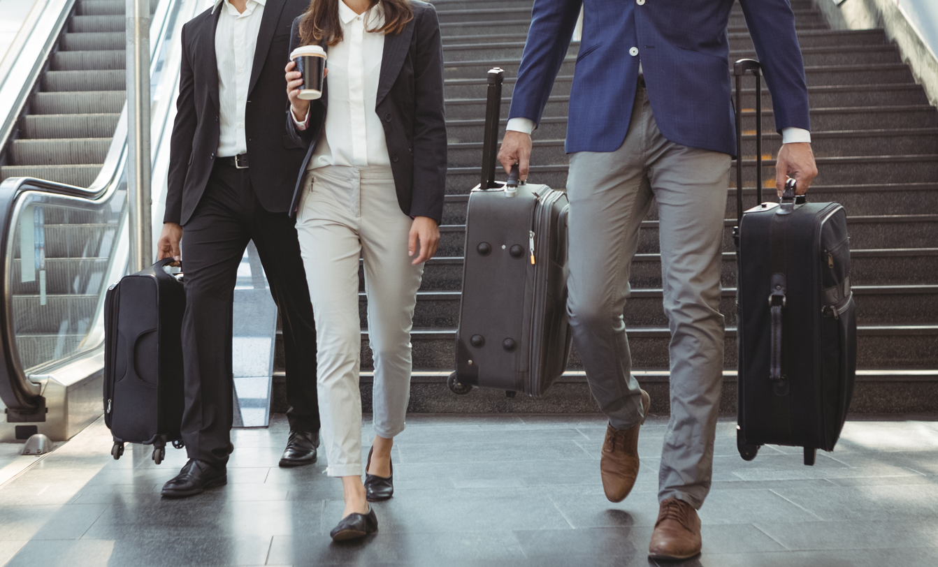 Tres profesionales vestidos formalmente caminando con maletas en un área de escaleras eléctricas, representando los viáticos como parte del ámbito empresarial.