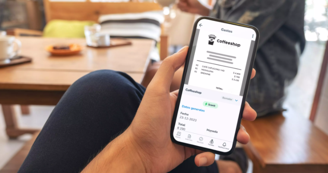 Persona revisando y registrando un gasto en su aplicación móvil desde una cafetería, con detalle del consumo en pantalla y ambiente relajado al fondo, ejemplificando cómo gestionar viáticos o gastos personales desde cualquier lugar.