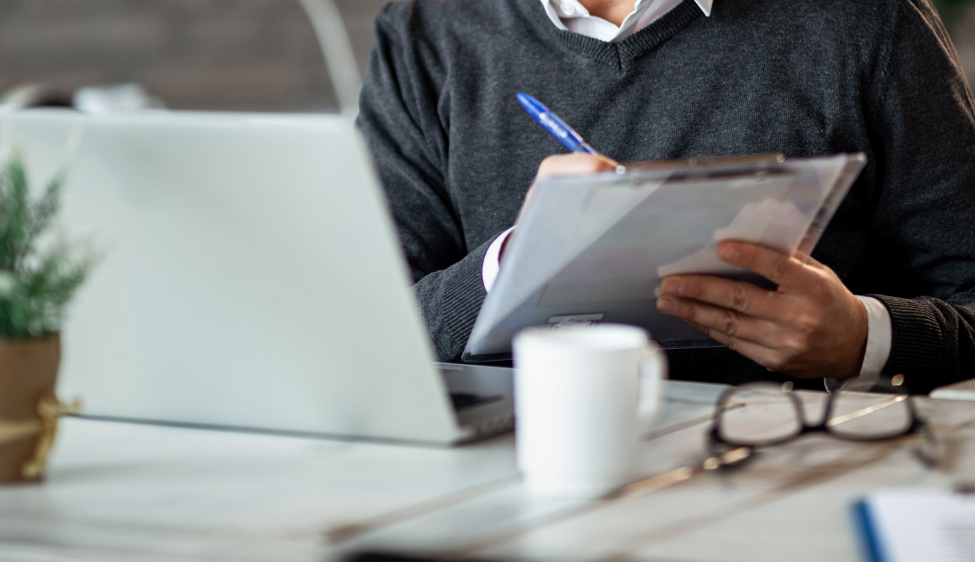 Hombre con suéter gris oscuro revisando información en un portapapeles mientras trabaja frente a una laptop en un ambiente de oficina, representando la revisión de gastos operativos.