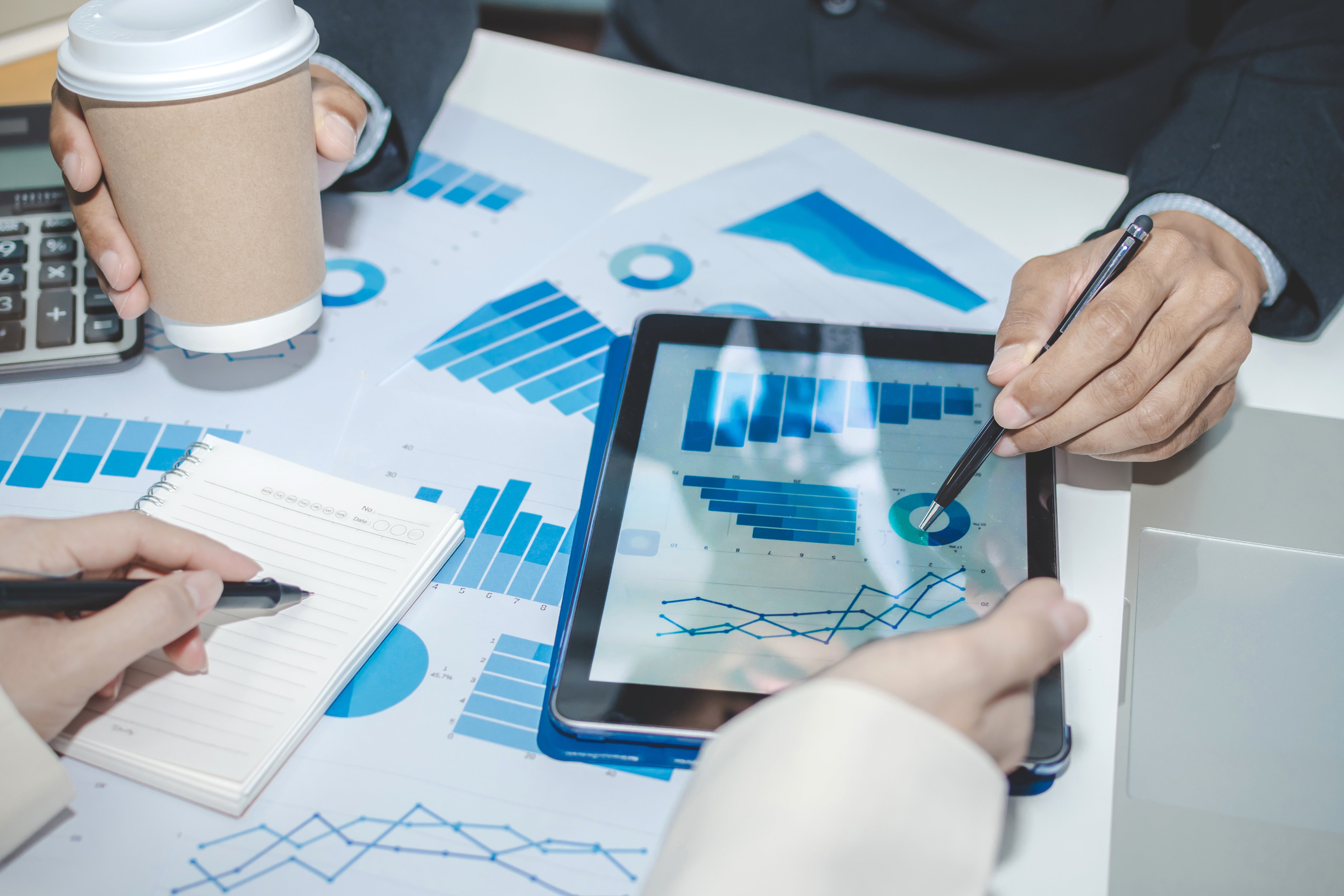 Varios profesionales trabajando juntos, revisando gráficos y datos de negocio en una tablet y tomando notas, con gráficos de barras y pastel visibles sobre la mesa.