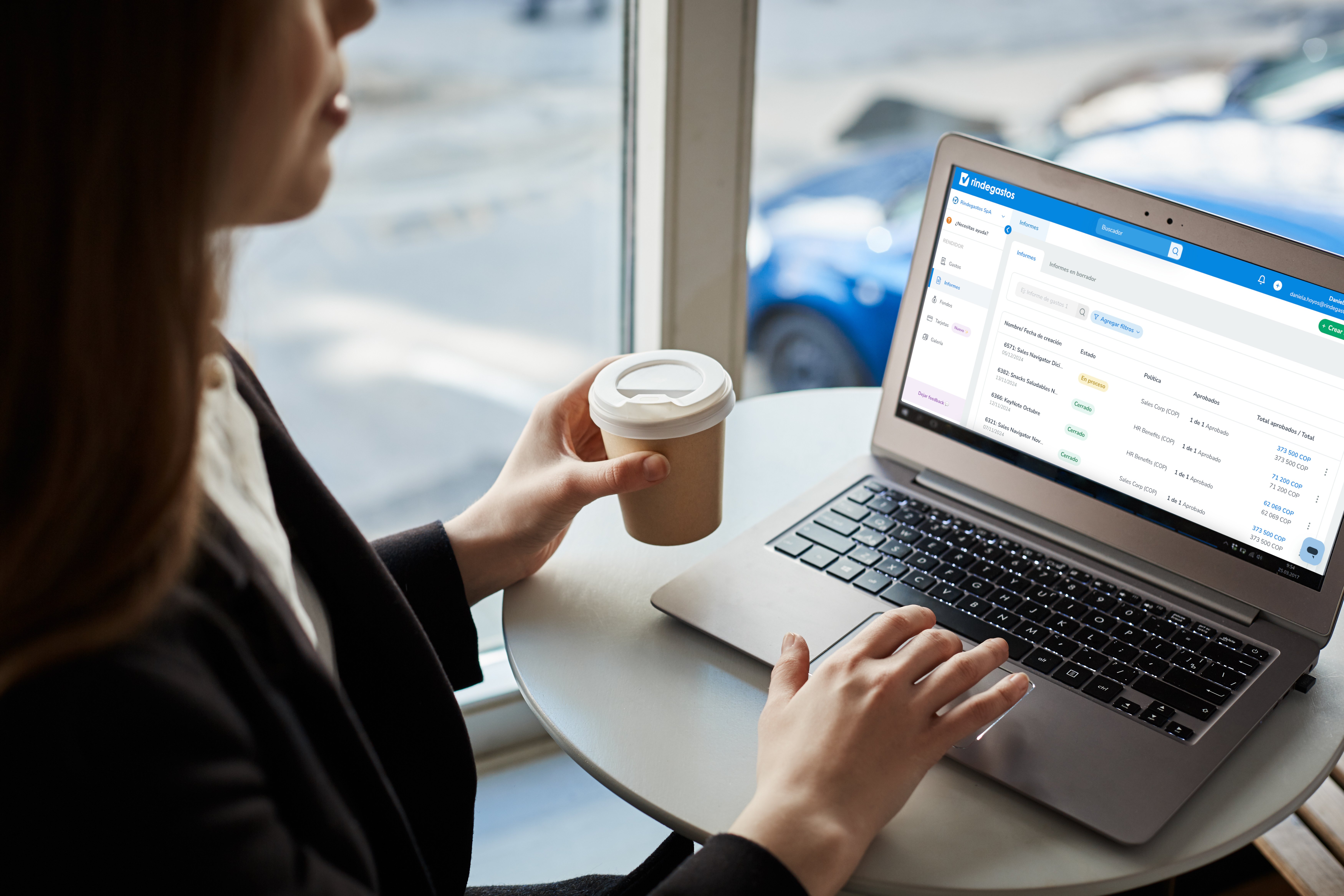 Mujer trabajando en una laptop en una cafetería, con la pantalla mostrando una interfaz de Rindegastos para gestionar gastos de viaje.