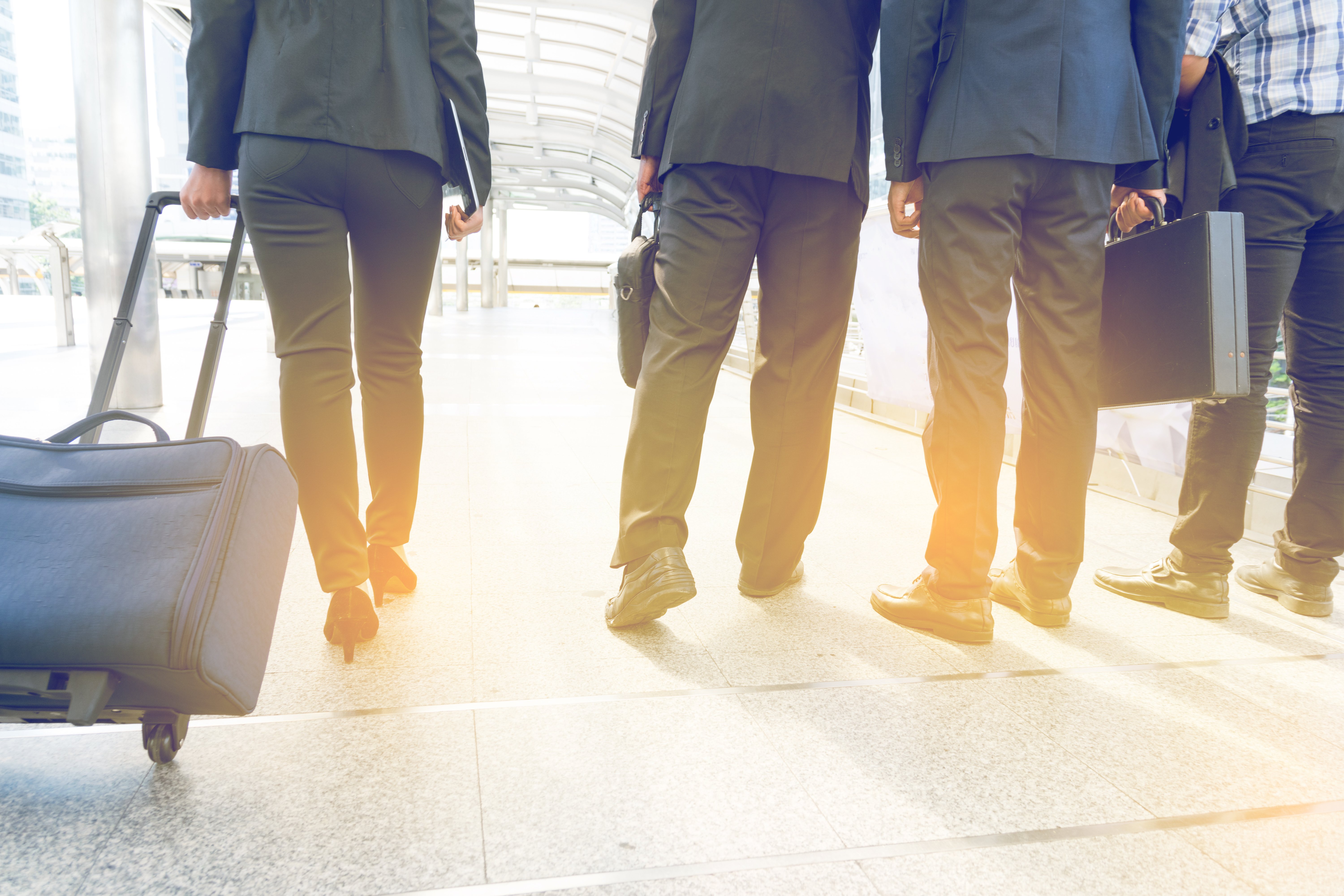 Cuatro personas de negocios caminando por una terminal de aeropuerto, llevando maletas y carpetas, en dirección hacia un destino.