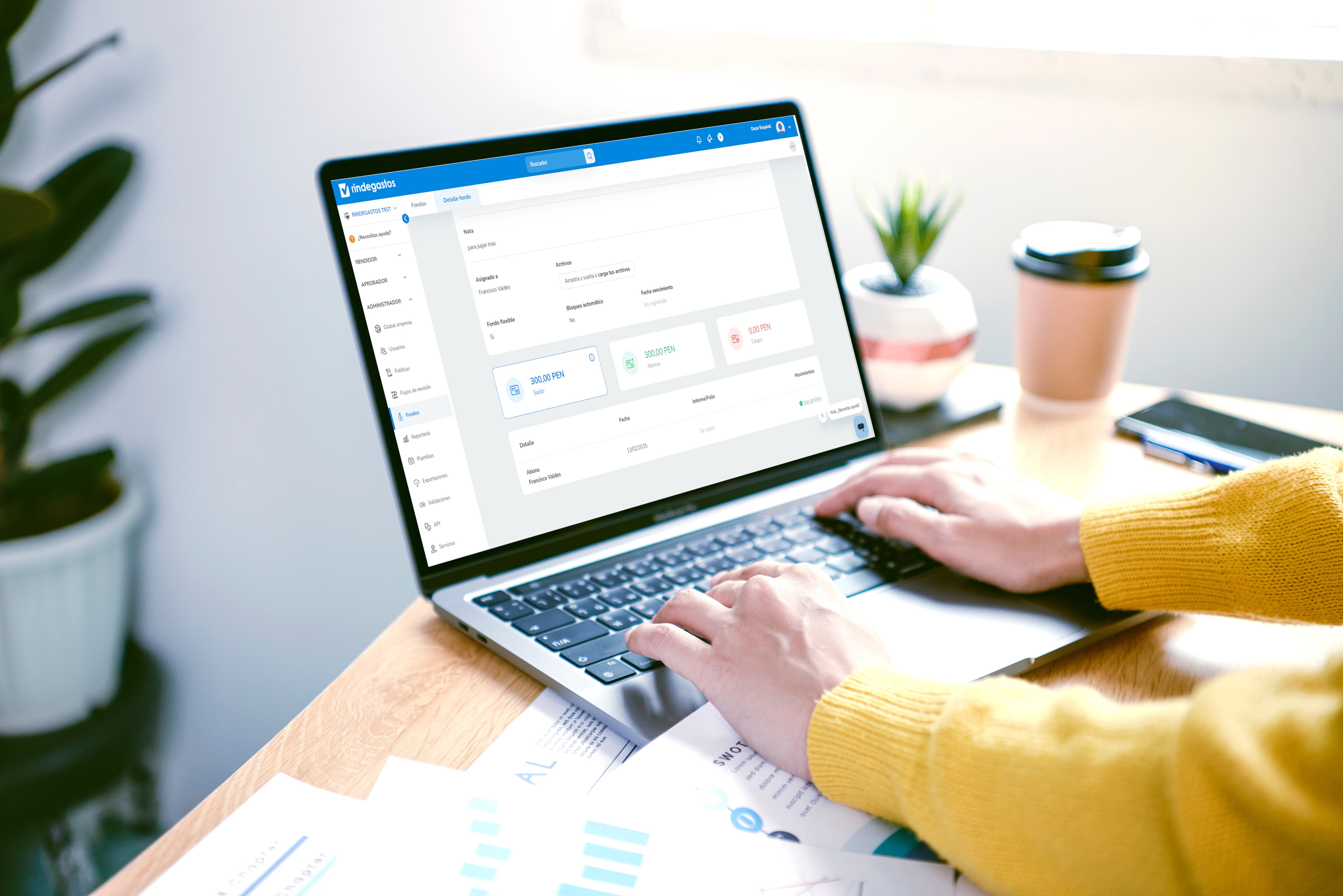Persona utilizando una computadora portátil en un escritorio, con una interfaz de software de gestión de gastos visible en la pantalla, junto a una taza y papelería.