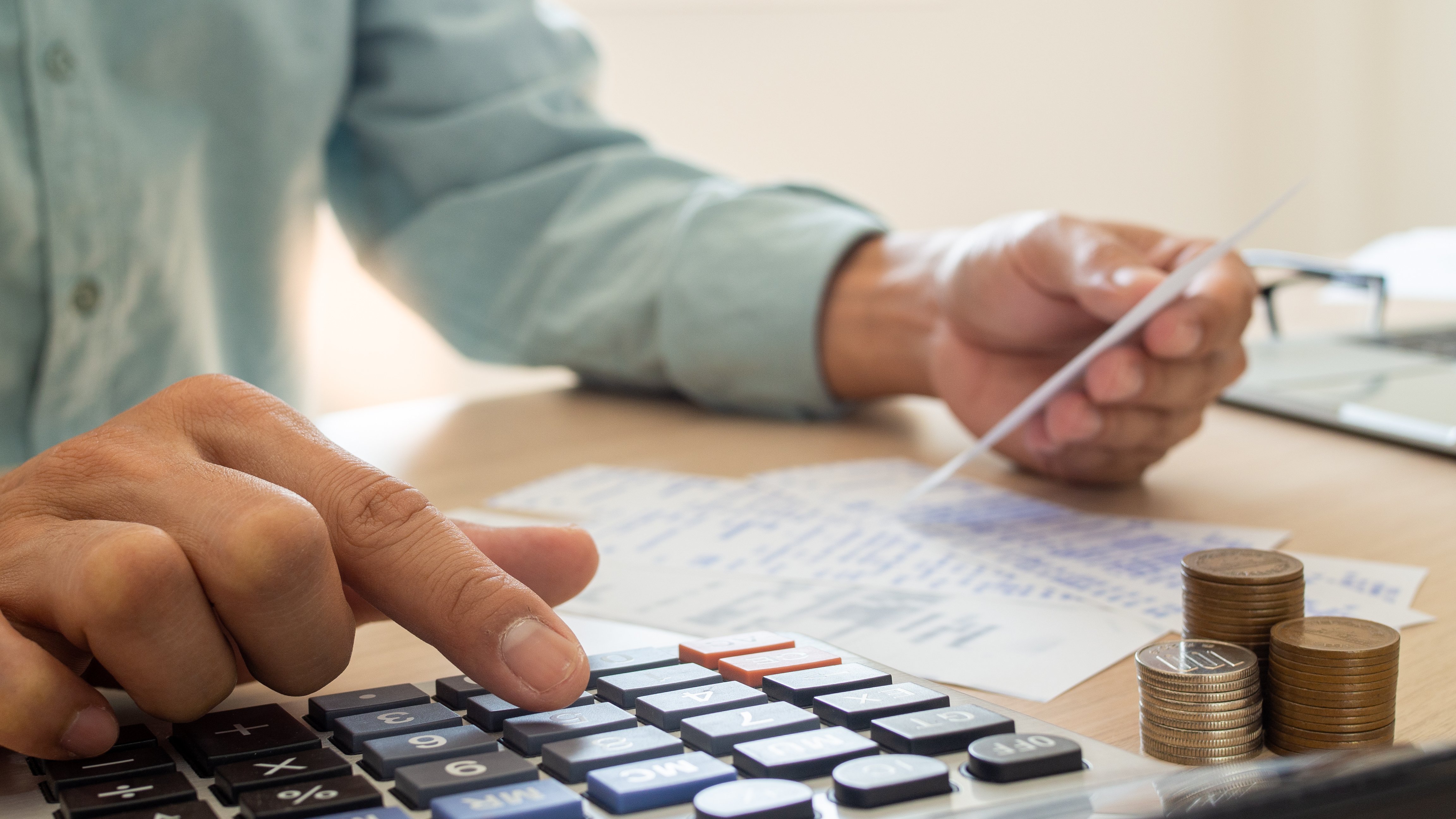 Persona utilizando una calculadora y sosteniendo una tarjeta mientras revisa documentos financieros, con pilas de monedas apiladas al lado, representando el análisis de gastos, pagos y presupuesto.