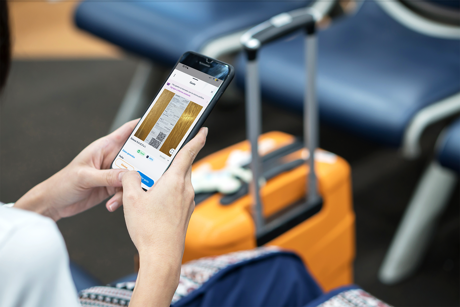 Viajera utilizando la aplicación móvil de Rindegastos para escanear documentos mientras espera su vuelo en aeropuerto, con maleta de cabina.