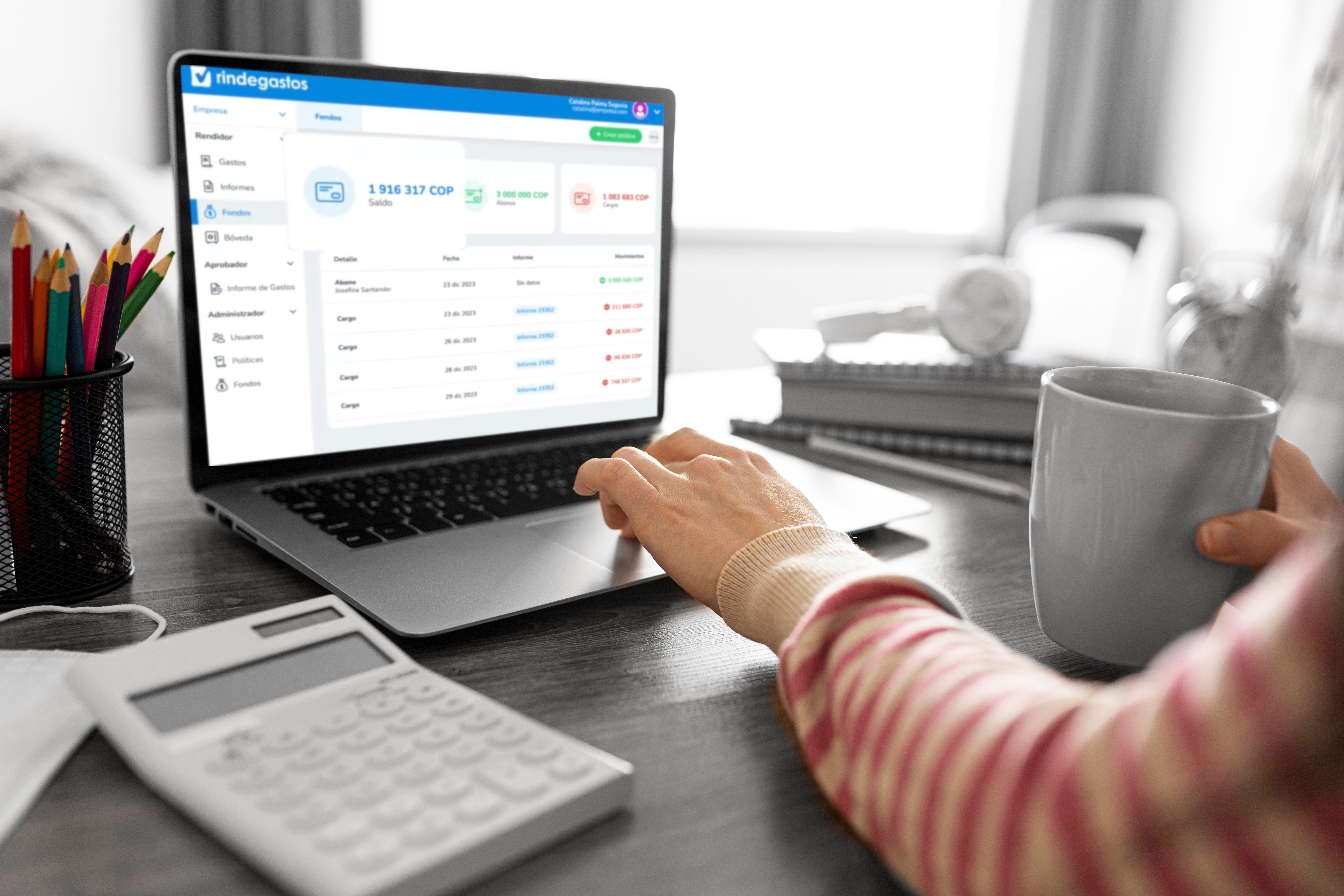 Pantalla de una computadora portátil mostrando Rindegastos, con una calculadora, una taza de café y lápices sobre el escritorio