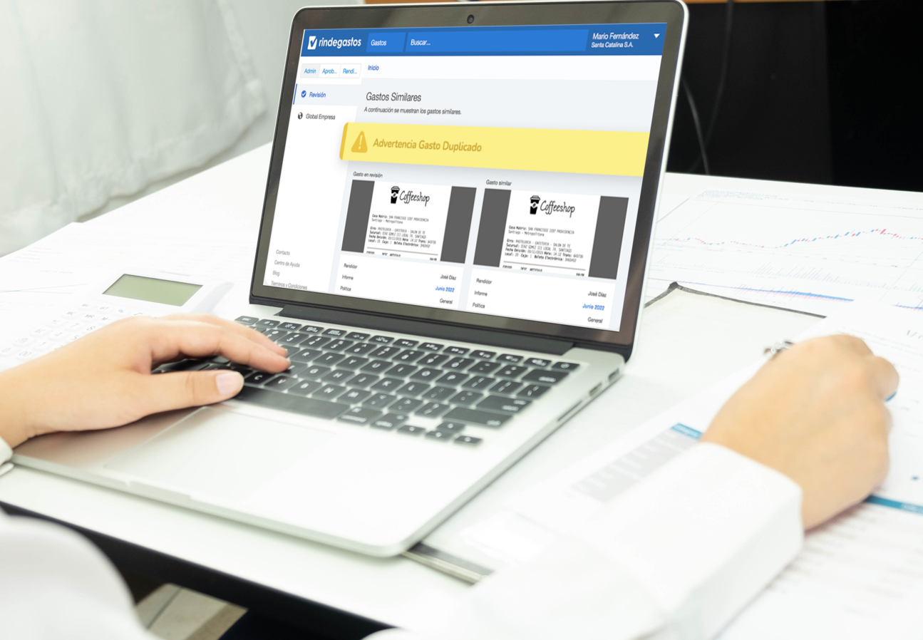 Persona utilizando la plataforma Rindegastos en su laptop, con una advertencia de "Gasto Duplicado", junto a documentos con gráficos y una pequeña planta decorativa.