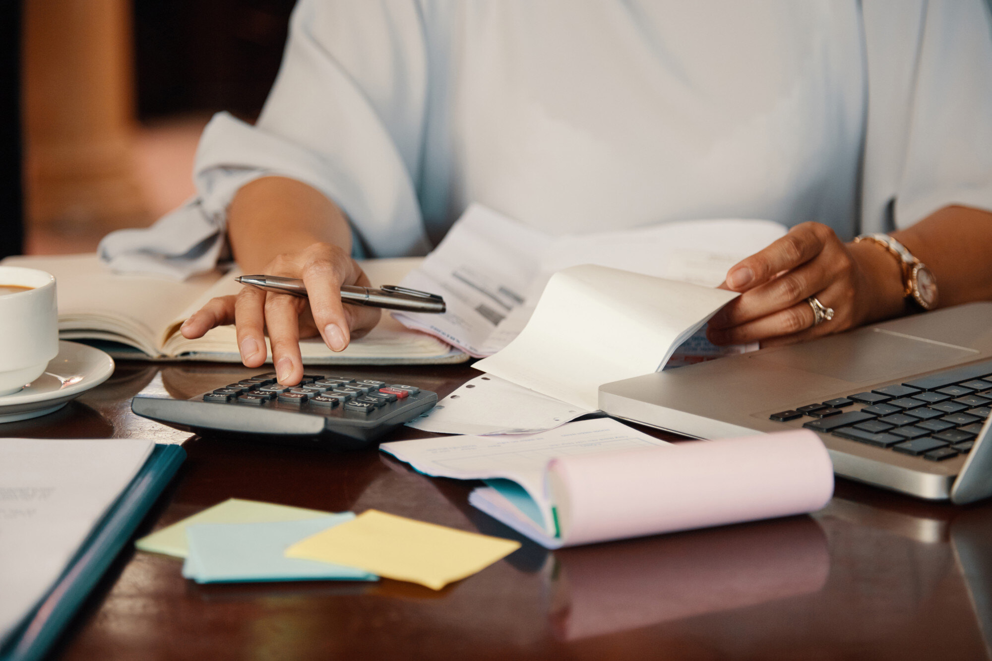 Mujer revisando documentos financieros con una calculadora y computadora portátil en un escritorio, acompañada de notas adhesivas y una taza de café.