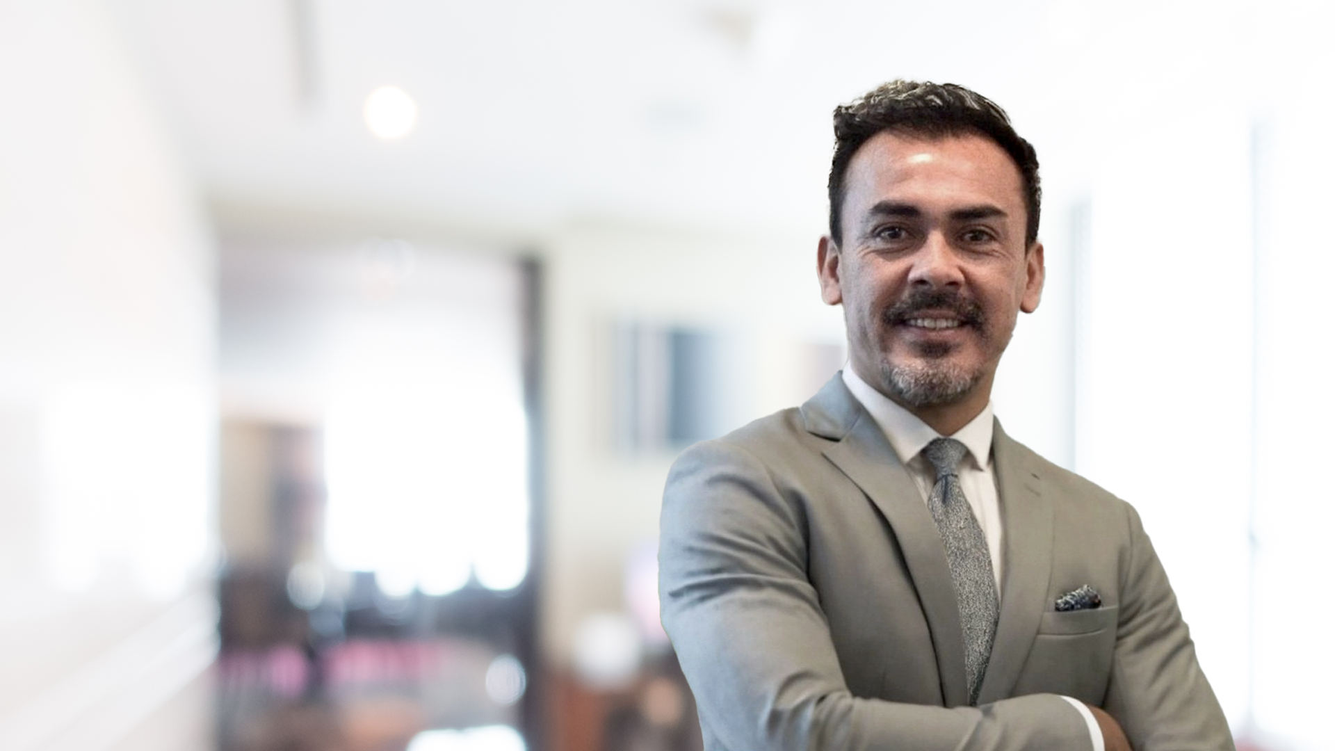 Hombre sonriente con traje gris y corbata, de pie con los brazos cruzados en una oficina moderna, representando liderazgo y profesionalismo