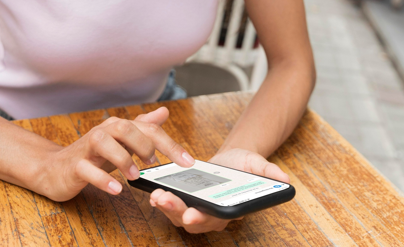  Una persona sosteniendo un teléfono móvil mientras usa WhatsApp en un entorno casual, como una cafetería. La pantalla del teléfono muestra una conversación en la que se envía la imagen de un recibo al chatbot, indicando la acción de registro de un gasto.