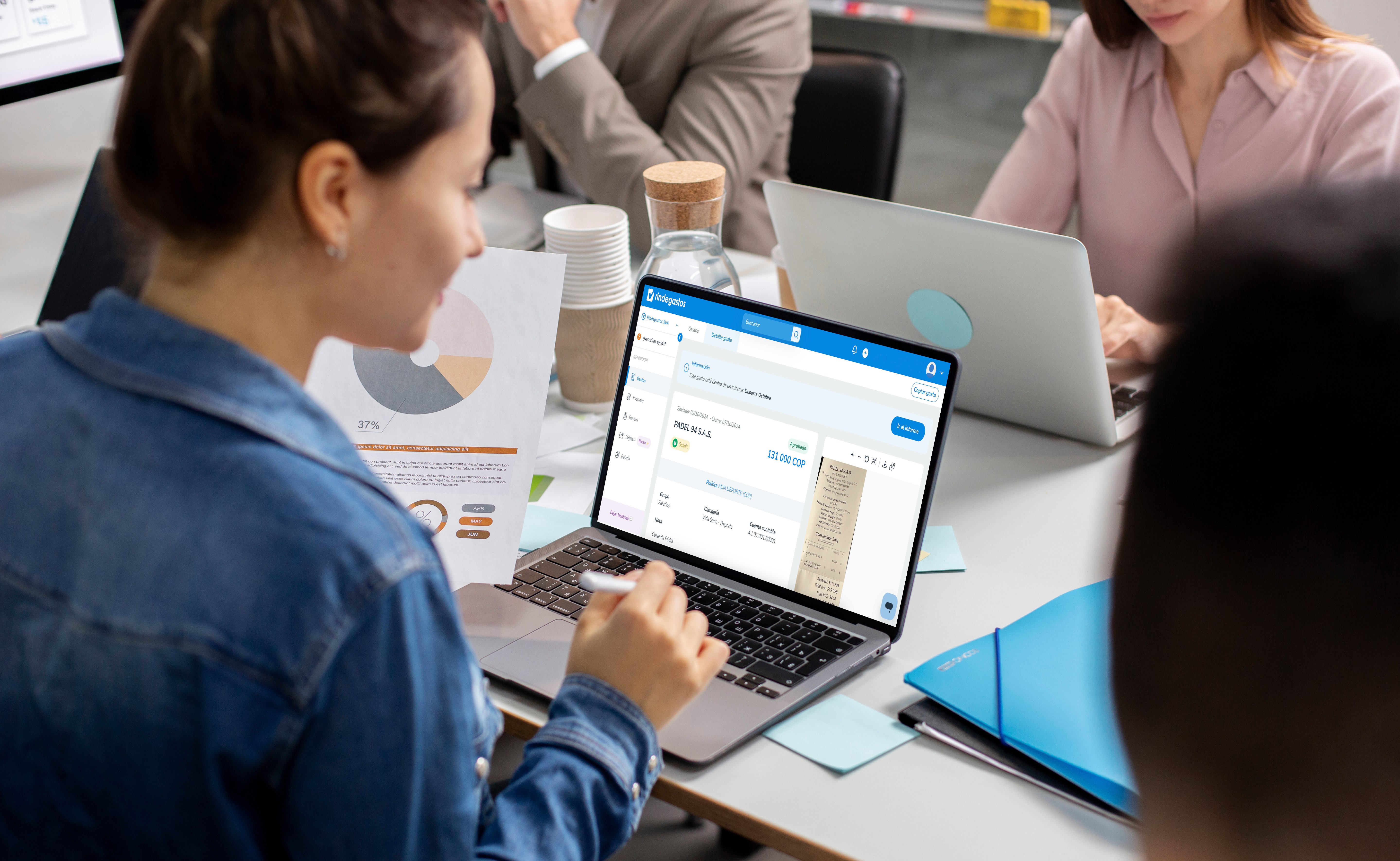 Mujer revisando la plataforma Rindegastos en su laptop durante una reunión de trabajo en oficina, con gráficos y documentos sobre la mesa.