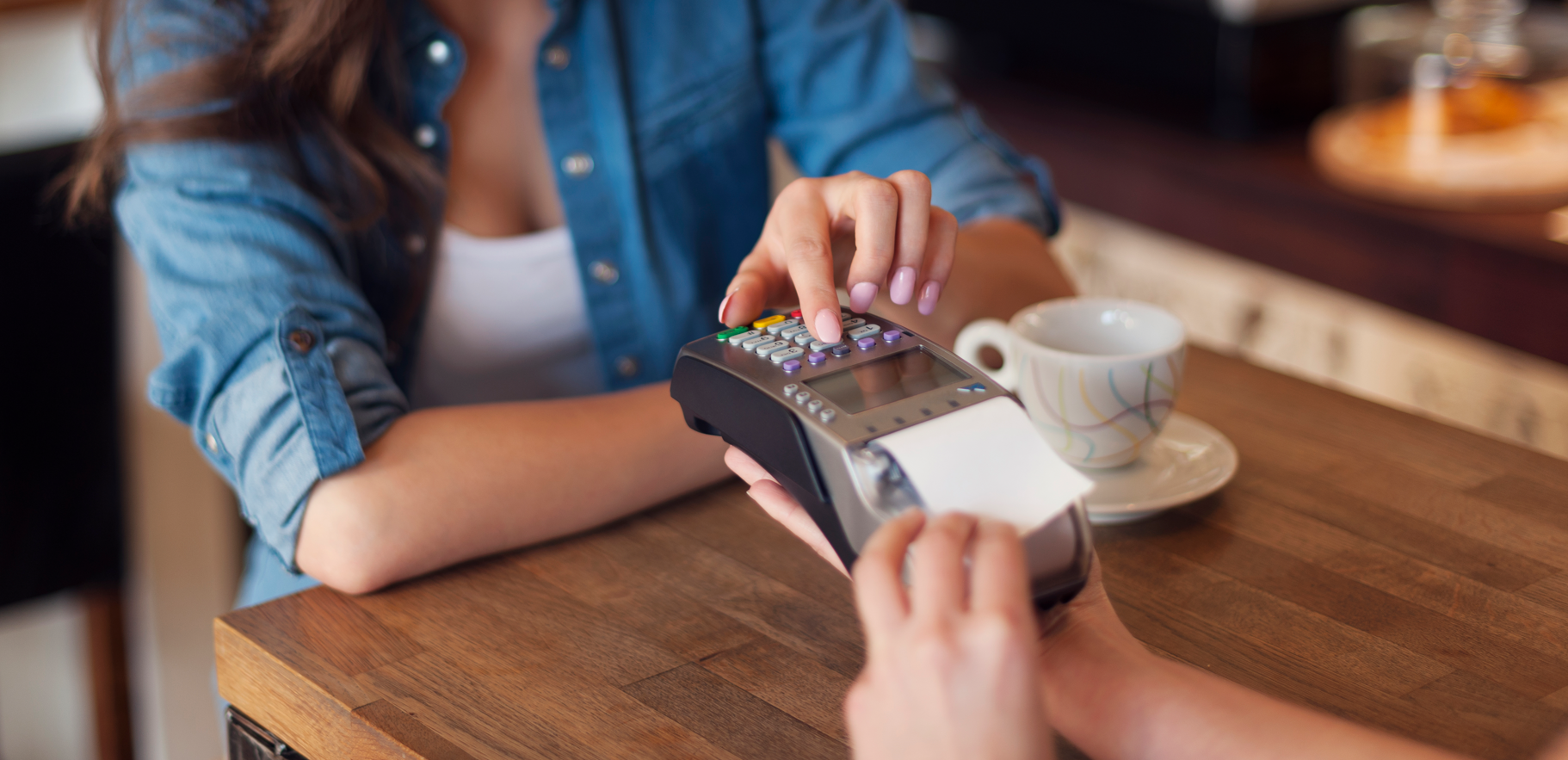 Mujer pagando en una cafetería con tarjeta de crédito en un lector de tarjetas de pago, con una taza de café en la mesa.