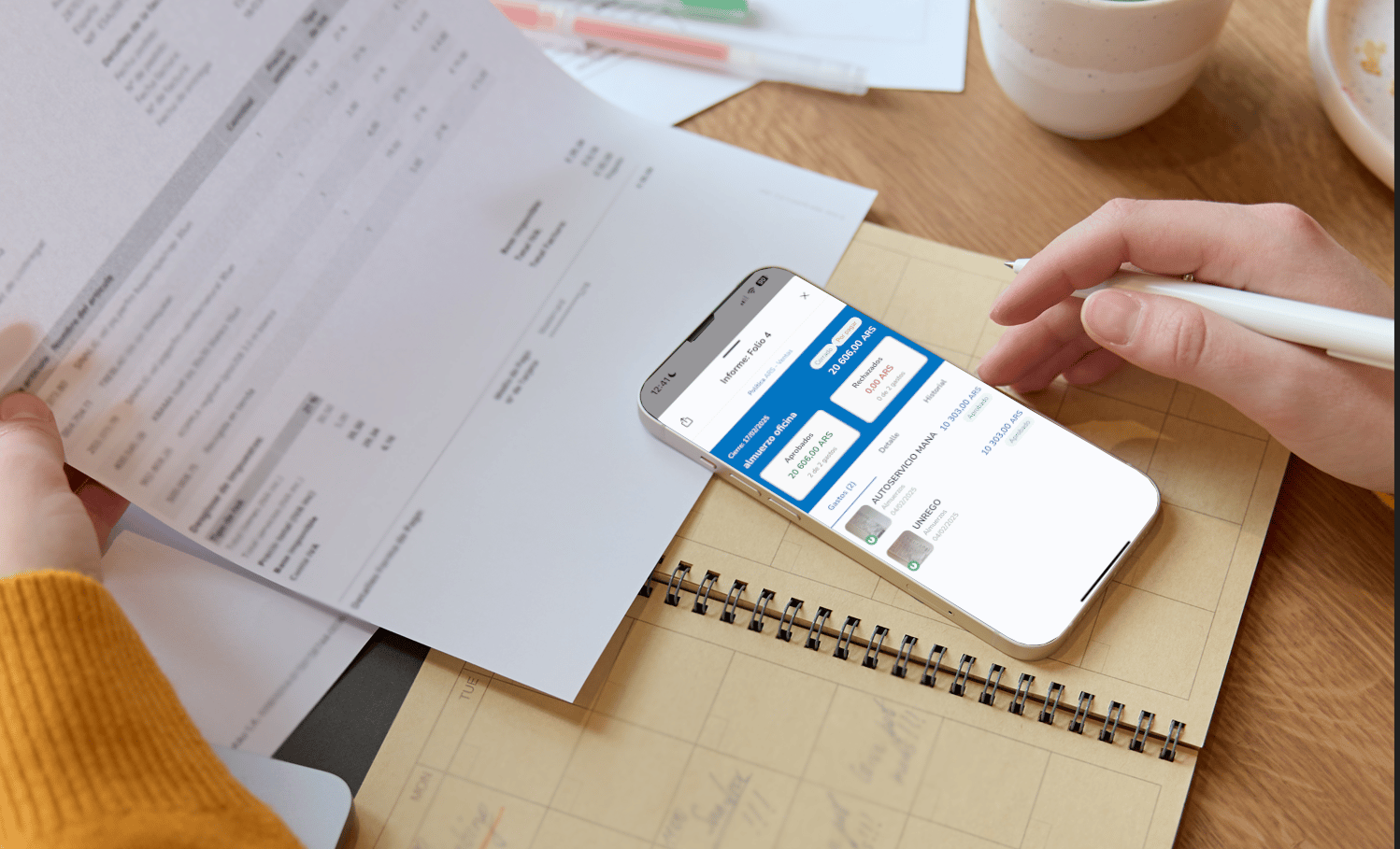 Persona revisando un recibo mientras usa un teléfono móvil con una aplicación de gestión de gastos visible en la pantalla, con documentos y una agenda en la mesa.