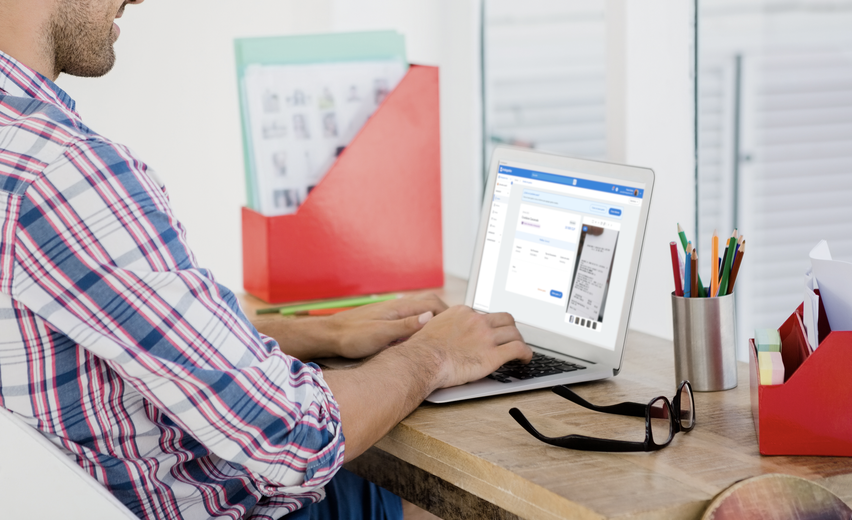 "Hombre con camisa de cuadros sentado en un escritorio, usando una laptop que muestra la interfaz de Rindegastos con detalles de recibos y aprobaciones, rodeado de carpetas y útiles de oficina.