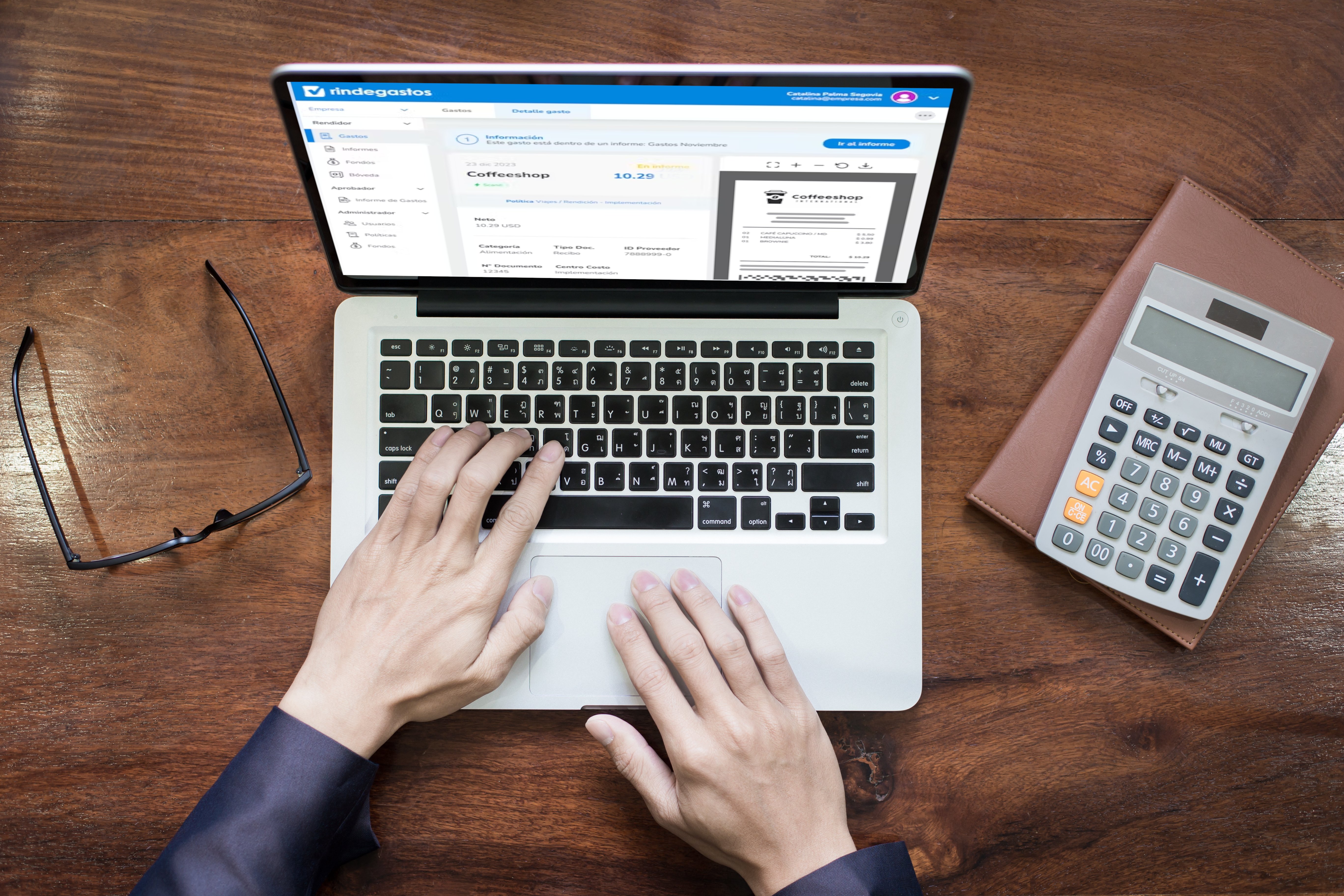 Vista superior de manos trabajando en una laptop con la plataforma Rindegastos abierta, acompañada de una calculadora y lentes sobre una mesa de madera