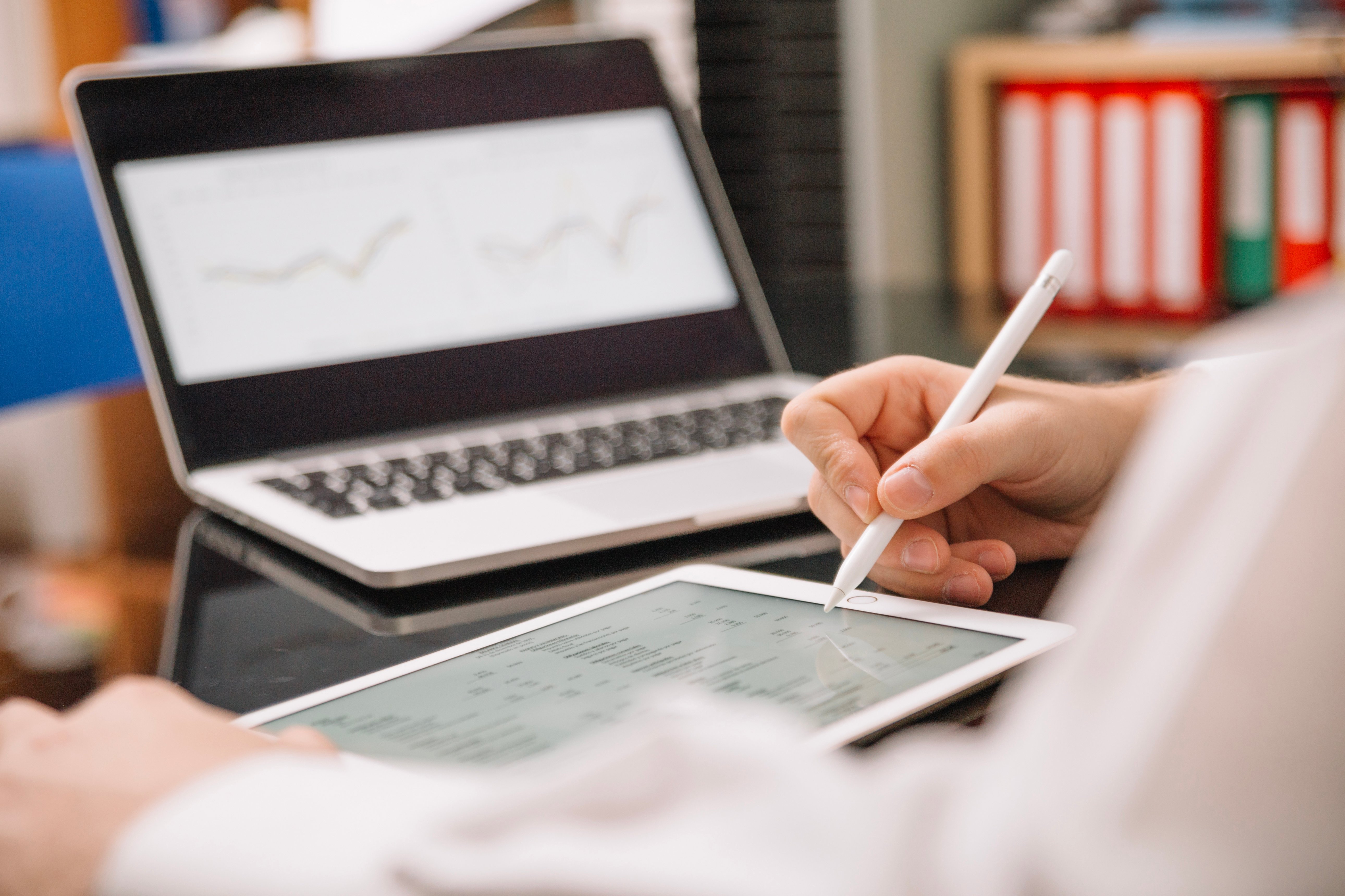 Persona usando un lápiz digital para interactuar con una tablet, mientras analiza datos financieros, con una laptop mostrando gráficos de líneas al fondo.