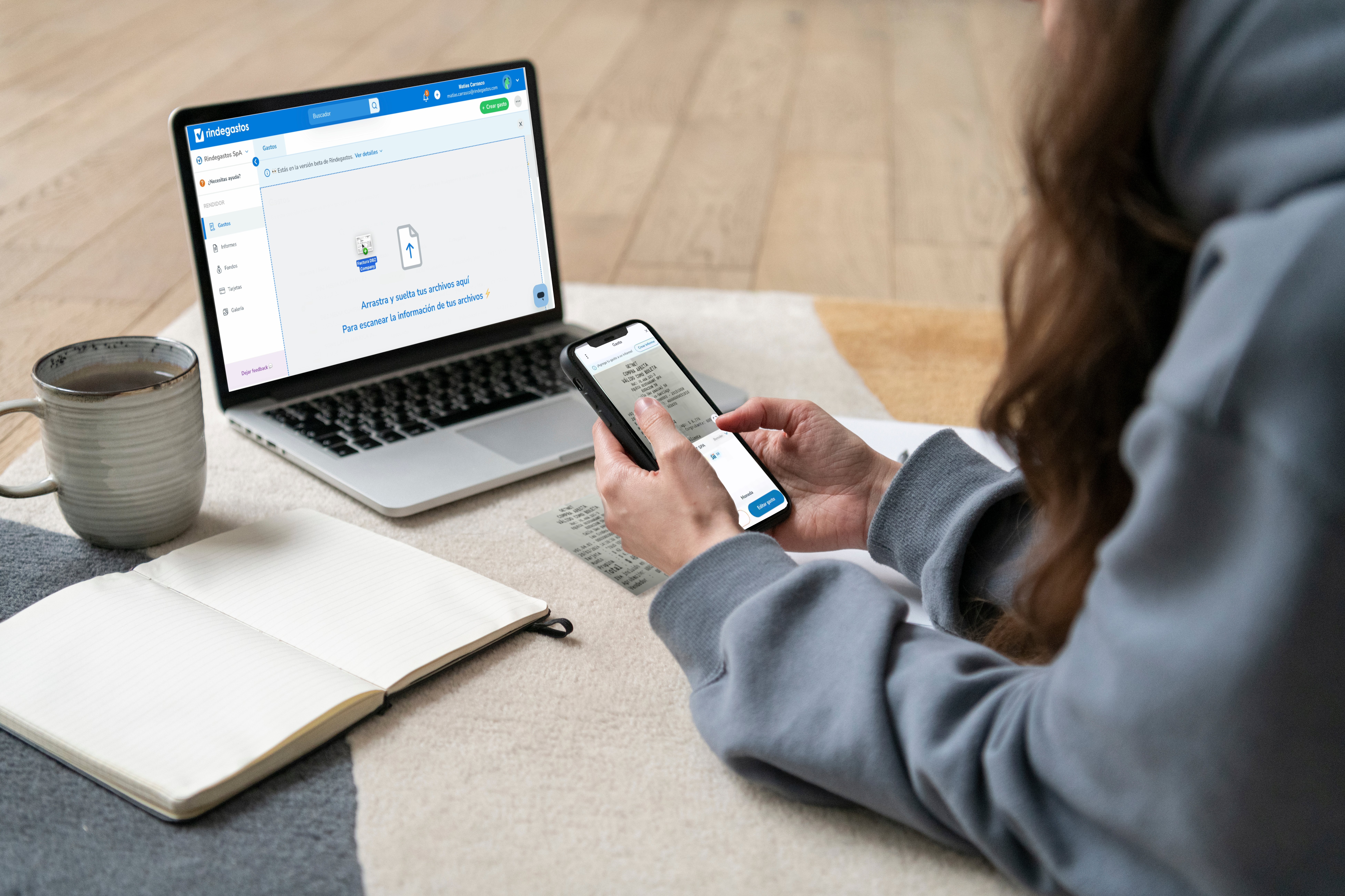 Mujer sentada frente a una laptop, escaneando una boleta smartphone. La laptop muestra una interfaz de de Rindegastos, mientras la mujer tiene una taza de café y un cuaderno abierto a su lado.